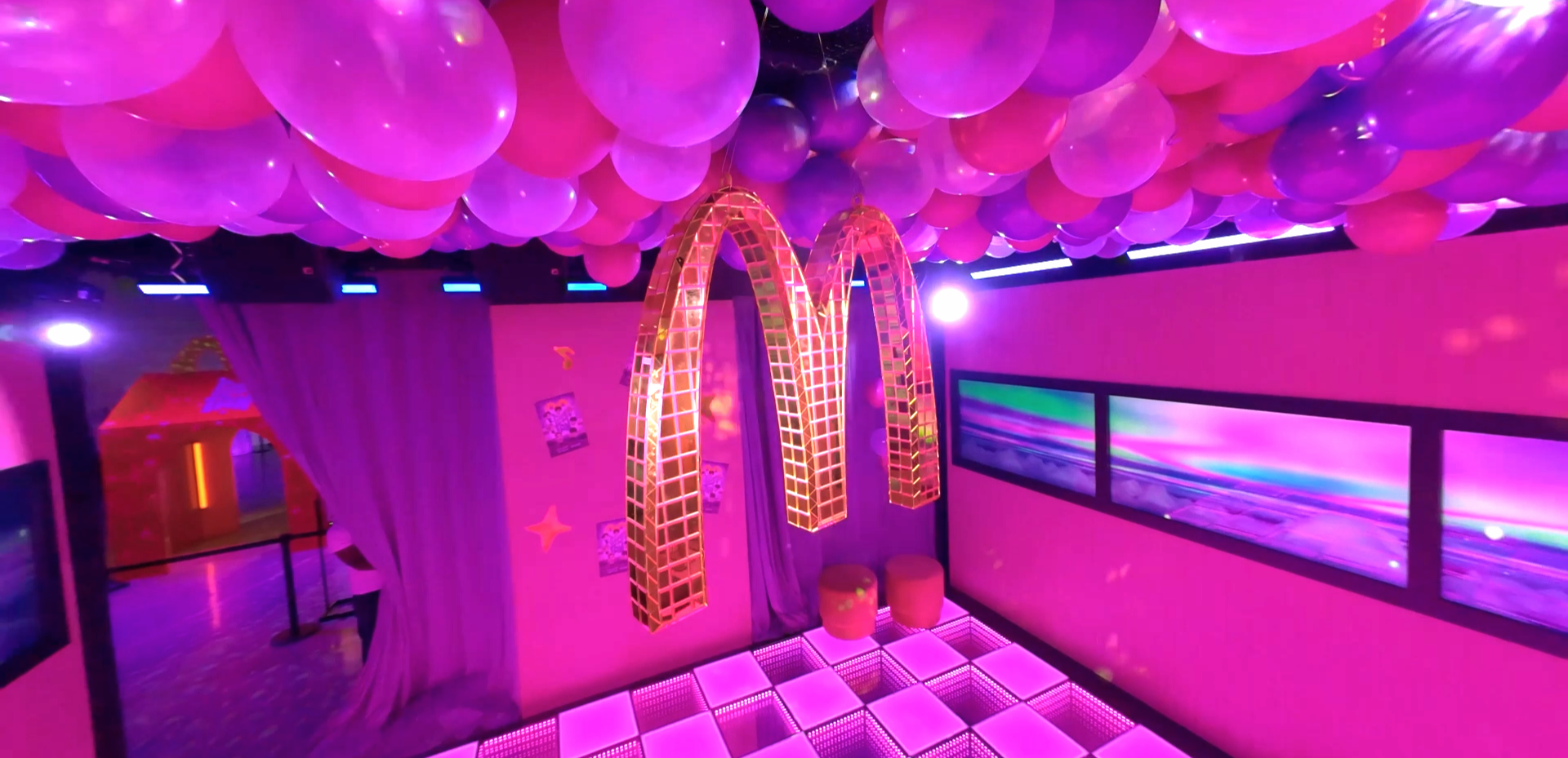Room decorated with pink, purple, and blue lighting, floating balloons on the ceiling, large illuminated golden arches, digital screens on the wall, and a checkered illuminated dance floor.