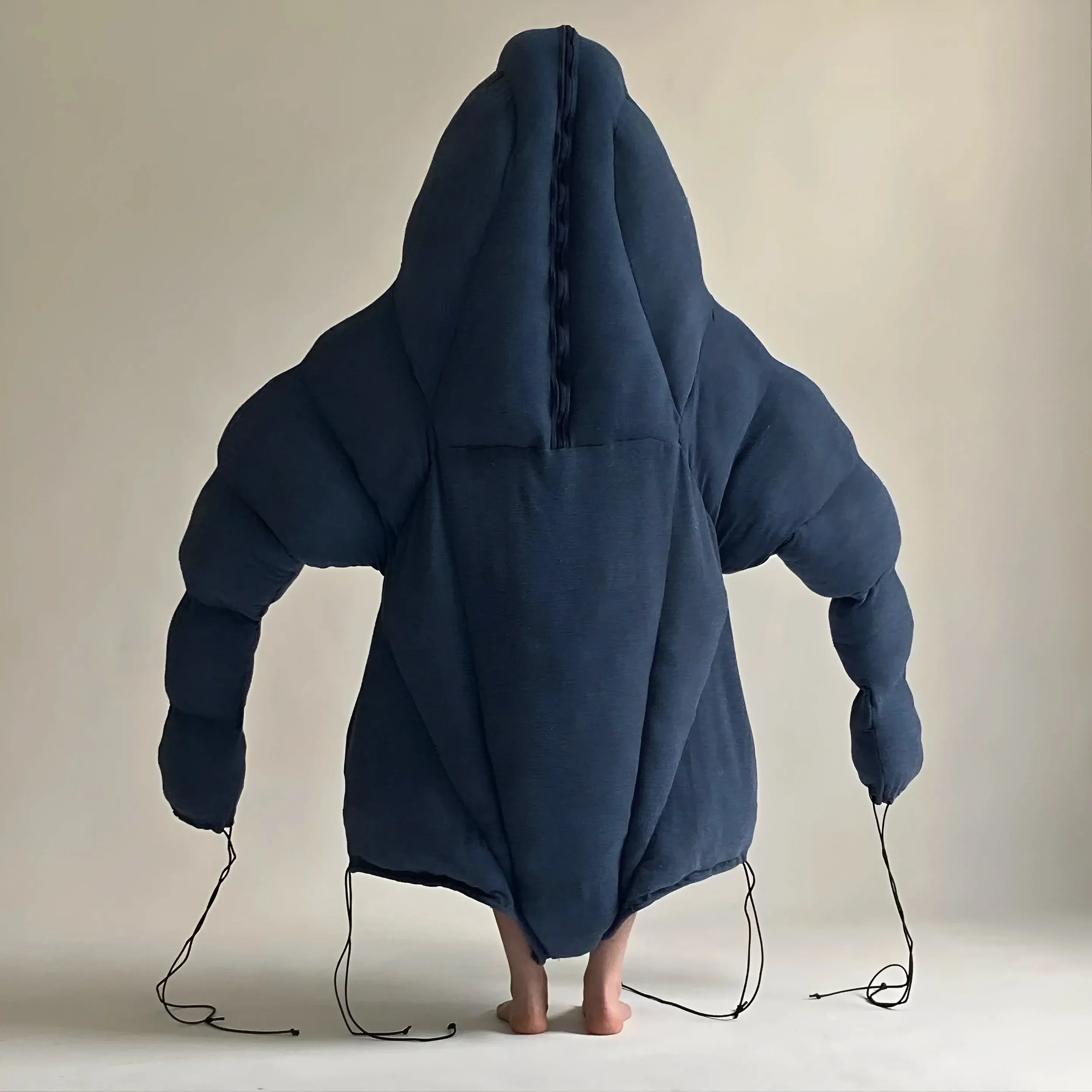 Person with a large navy blue padded jacket with a hood, standing barefoot against a plain white wall.