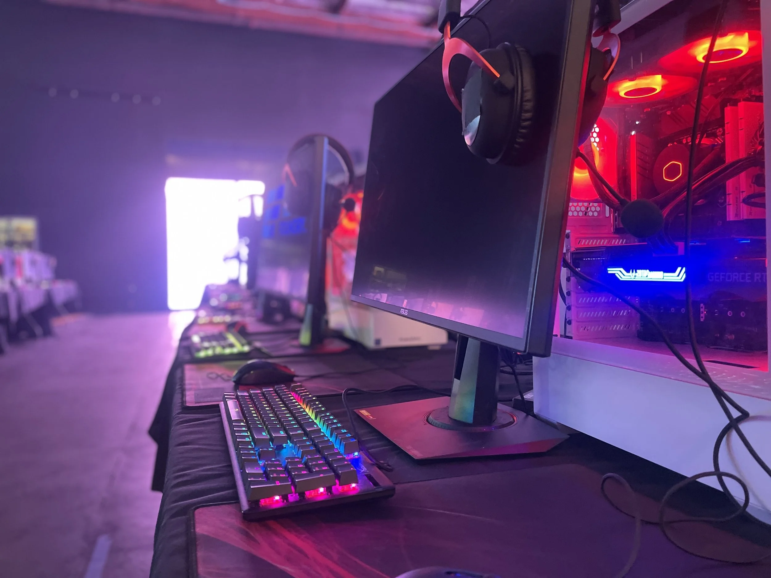 Row of gaming computers with monitors, keyboards, mice, and headphones, illuminated by colorful LED lights in a dark room.