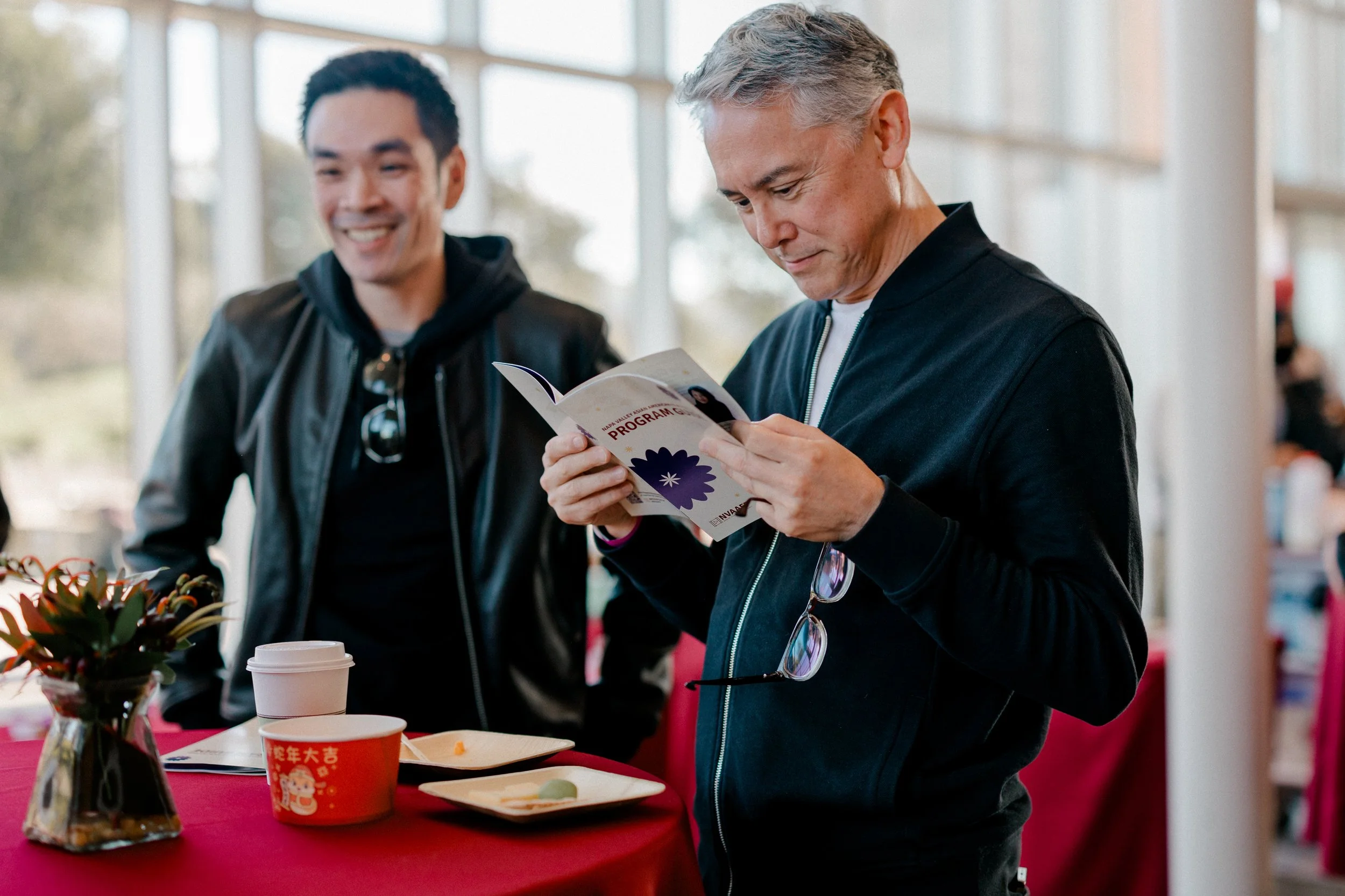 Two men stand at a table in a bright indoor space. One man, wearing a black jacket, is smiling with sunglasses hanging from his collar. The other man, with gray hair and wearing a dark zip-up jacket, is looking at a brochure or program he is holding. There are cups, plates, and a potted plant on the table.