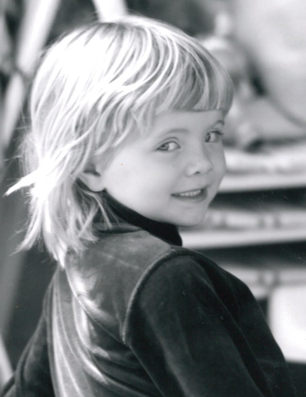 A young girl with light-colored hair smiling and looking over her shoulder in a black and white photo.