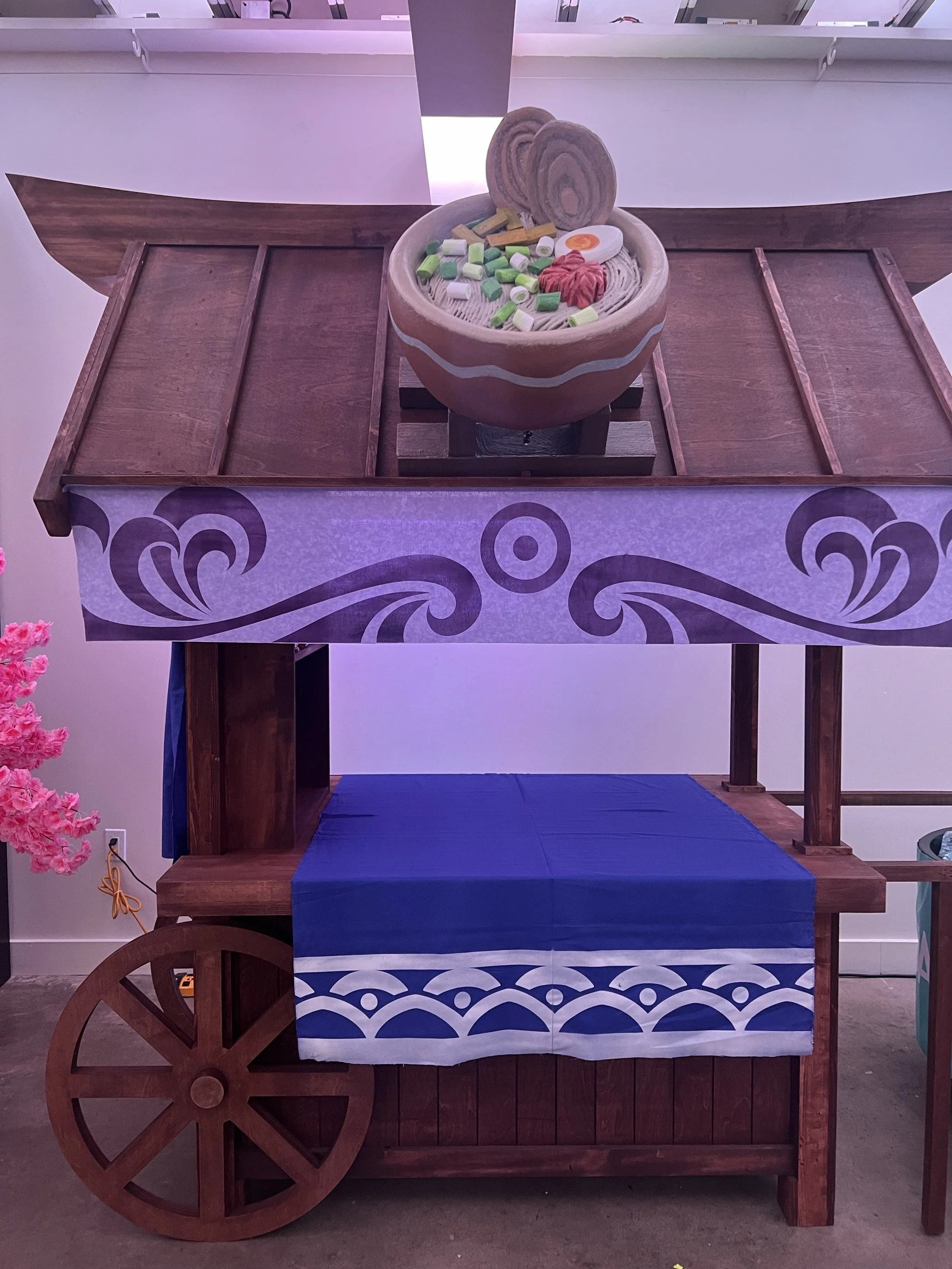 A wooden cart with a purple cloth and a large bowl on top, filled with plastic food toys resembling ramen with noodles, eggs, and vegetables, set in a room with white walls and pink lighting.