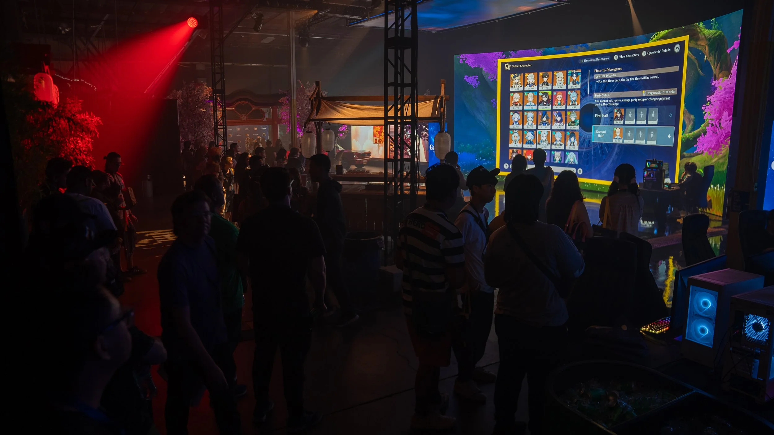 People at a gaming event with a large screen displaying a video game interface, illuminated by red and colorful lights, in a dimly lit, techy indoor venue