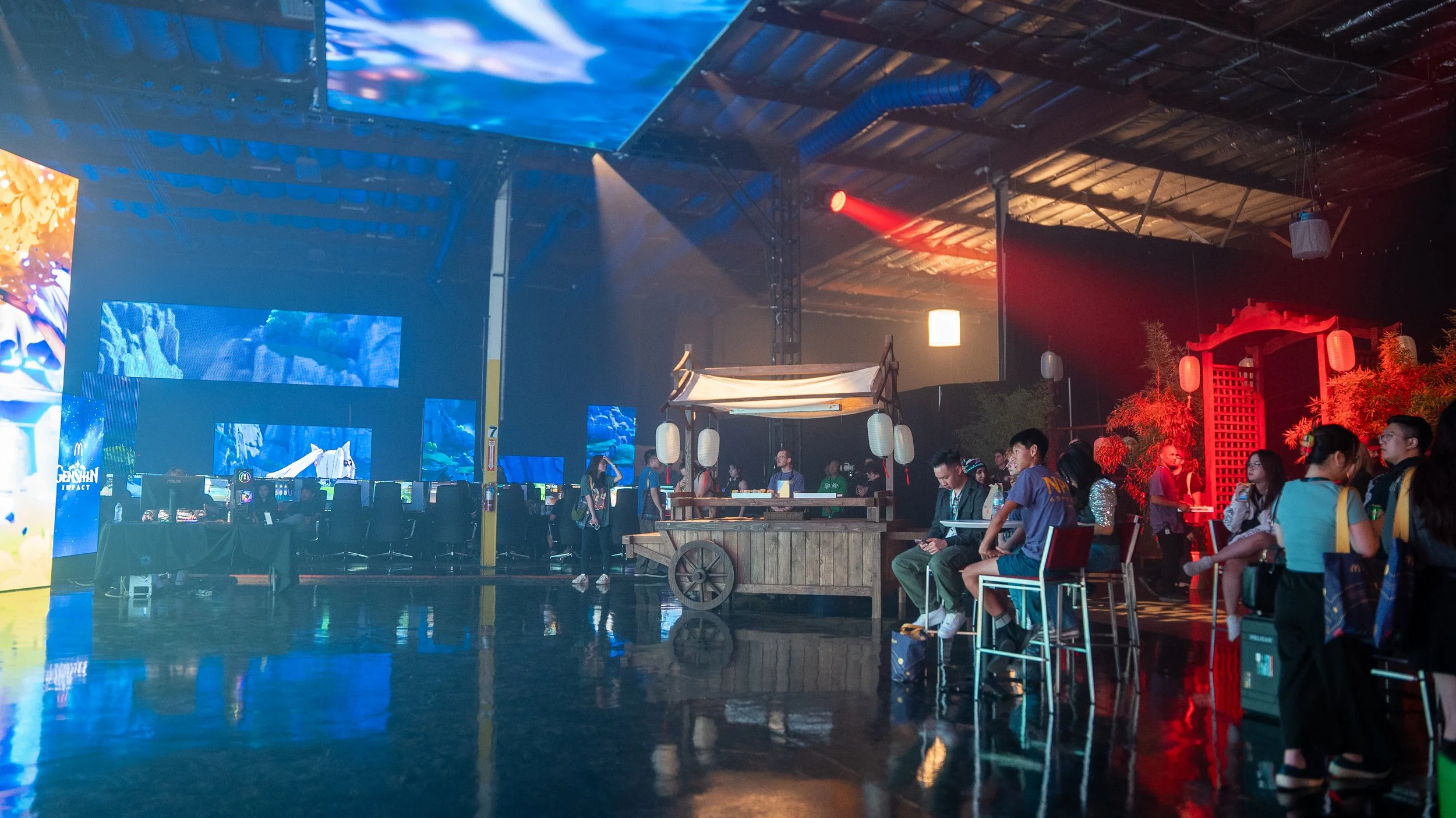 People at the Genshin LAN Party (indoor gaming event) with large screens, gaming stations, and Asian-themed decorations including a wooden cart, paper lanterns, and red lighting.
