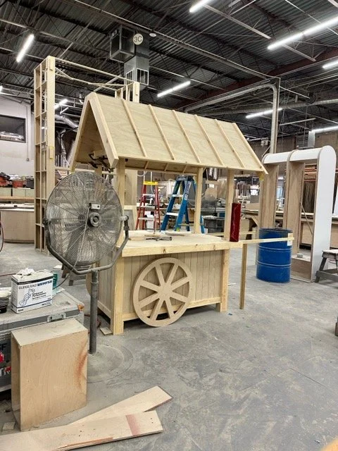 Wooden cart with a roof, resembling a small marketplace stall, in a woodworking workshop.