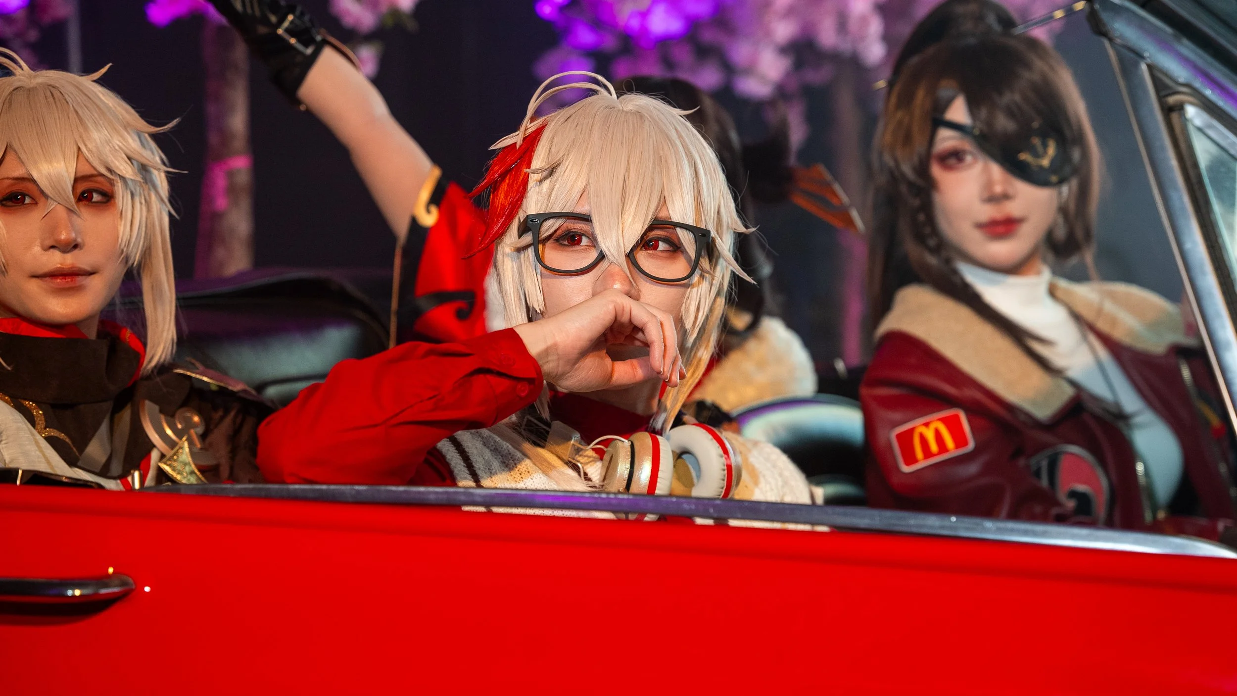 Three people in cosplay costumes sitting in a red vehicle, with one person wearing glasses and making a heart shape with their hand, with pink and purple lights and floral decorations in the background.