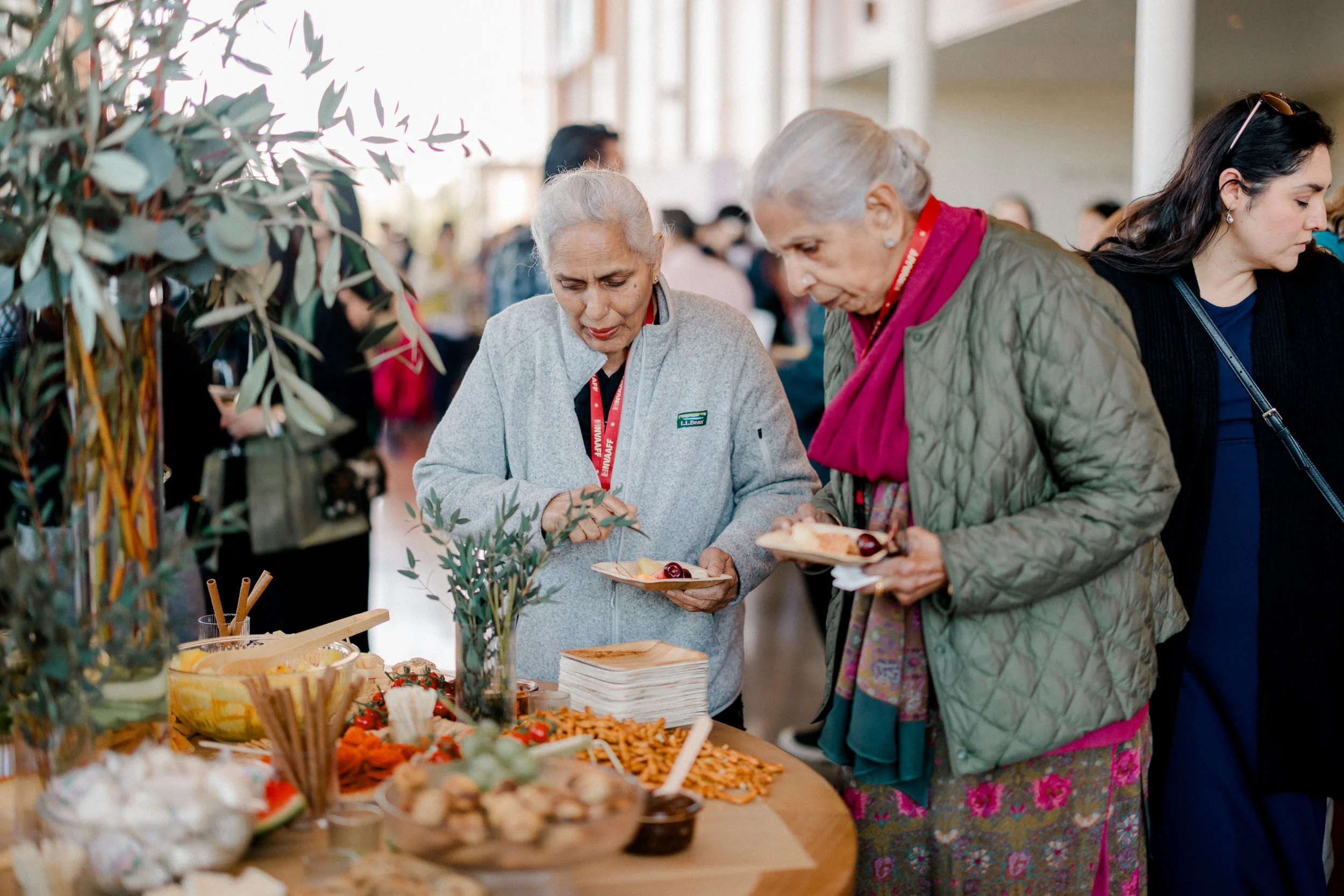 Two elderly women are at a food buffet, serving themselves from a variety of dishes on a wooden table. The table has salads, bread, and other foods, with a green plant centerpiece. The background shows a busy indoor event with other attendees.