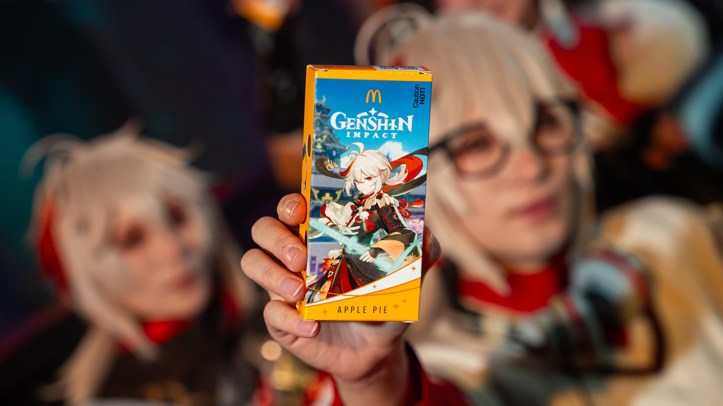 A cosplayer with white hair, glasses, and a colorful costume holding a McDonald's x Genshin Impact Apple Pie Box with an anime character illustration, while blurring the person's face in the background.