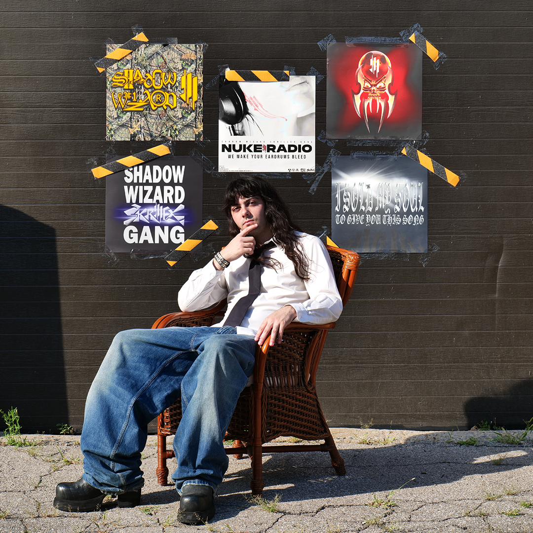 Person with long dark hair sitting in a wicker chair outdoors, wearing a white shirt, black tie, ripped jeans, and black boots, with a dark wooden wall behind them featuring six posters taped to it.