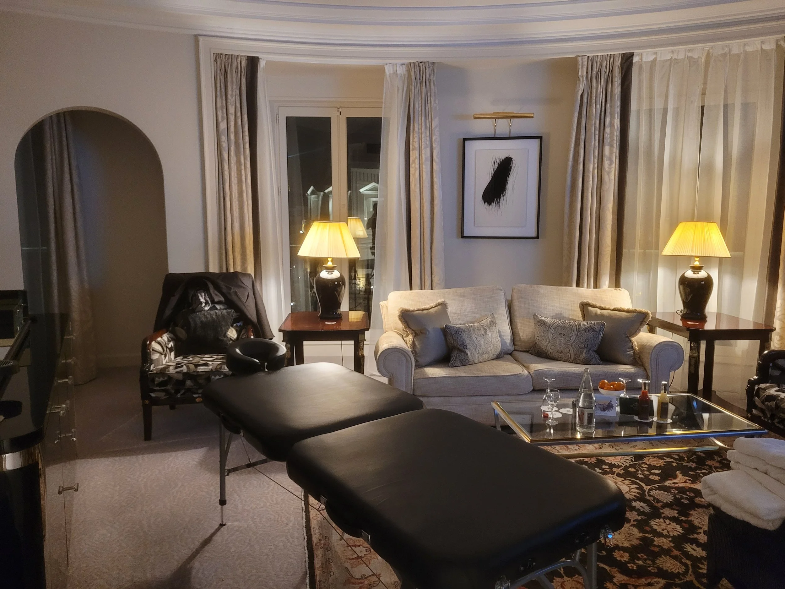 Salon élégant avec un sofa beige, deux tables d'appoint avec lampes, une table basse en verre avec des bouteilles et des verres, un fauteuil avec une couverture, une table de massage, de grandes fenêtres avec rideaux, décoration moderne et ambiance chaleureuse.