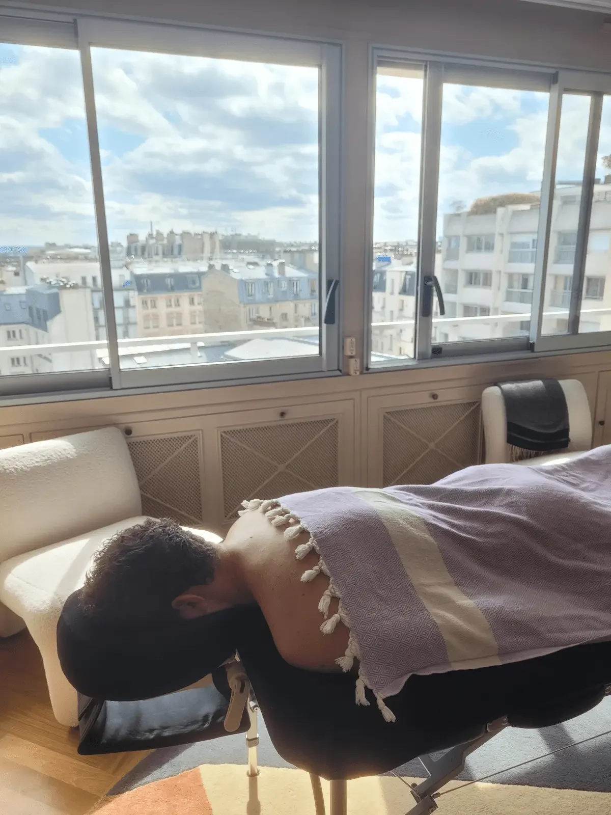 Une personne allongée sur une table de massage près d'une grande fenêtre donnant sur une vue urbaine avec des bâtiments modernes et un ciel partiellement nuageux, dans une pièce bien éclairée.