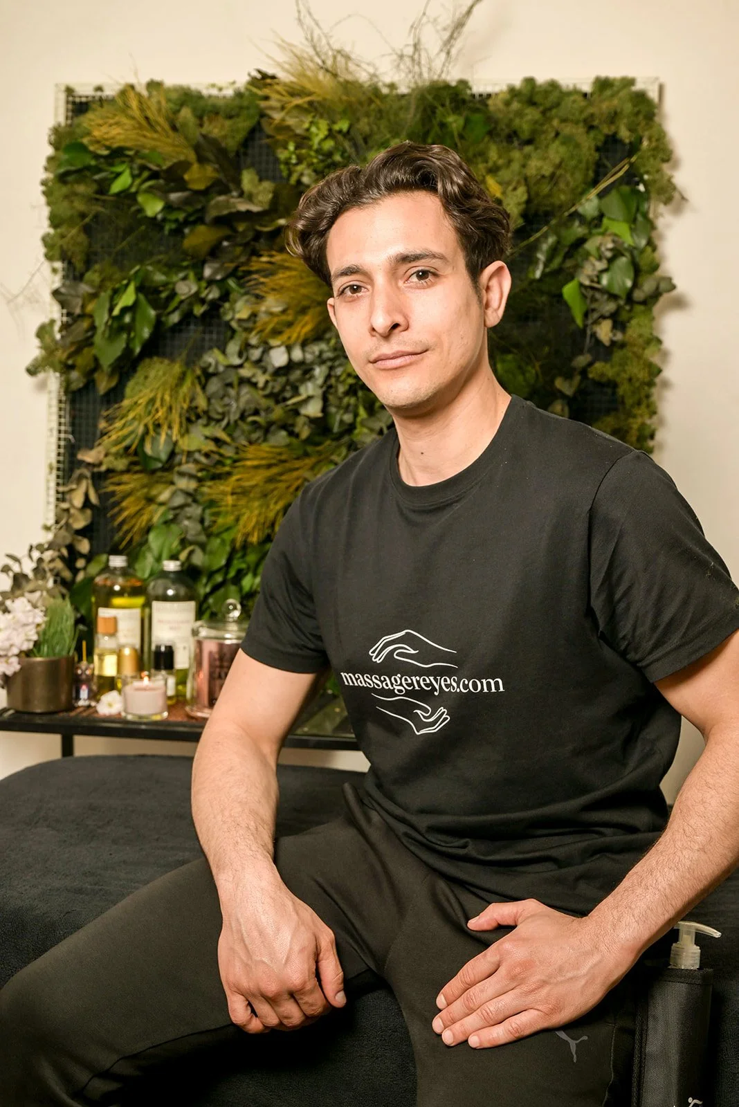 Un homme assis sur une table de massage, portant un t-shirt noir avec le texte 'massagereyes.com', en arrière-plan un mur végétal avec des plantes et des bouteilles de produits de massage, dans une salle de spa ou de massage.