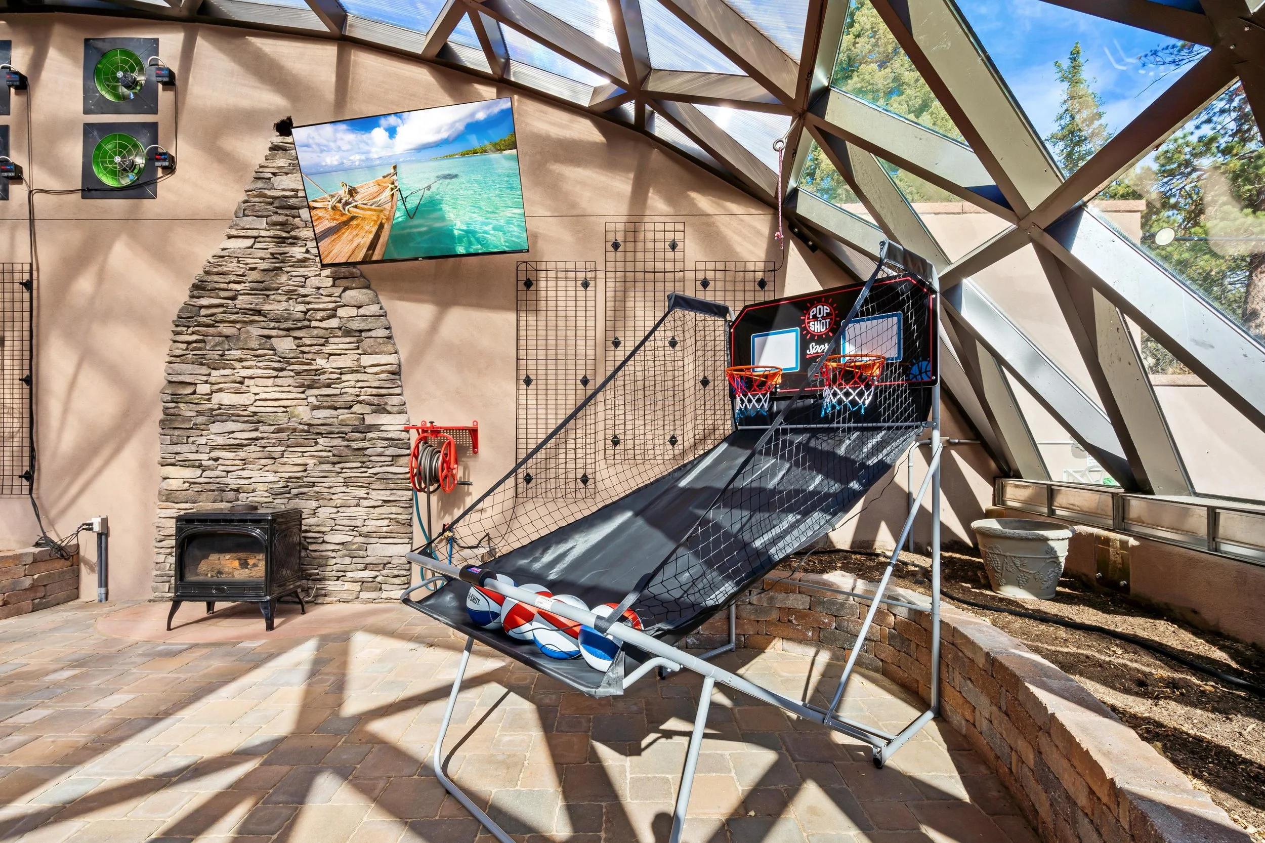 Indoor recreational area with a basketball arcade game, a stone fireplace, a wall-mounted TV showing a tropical beach scene, and large glass windows letting in natural light.