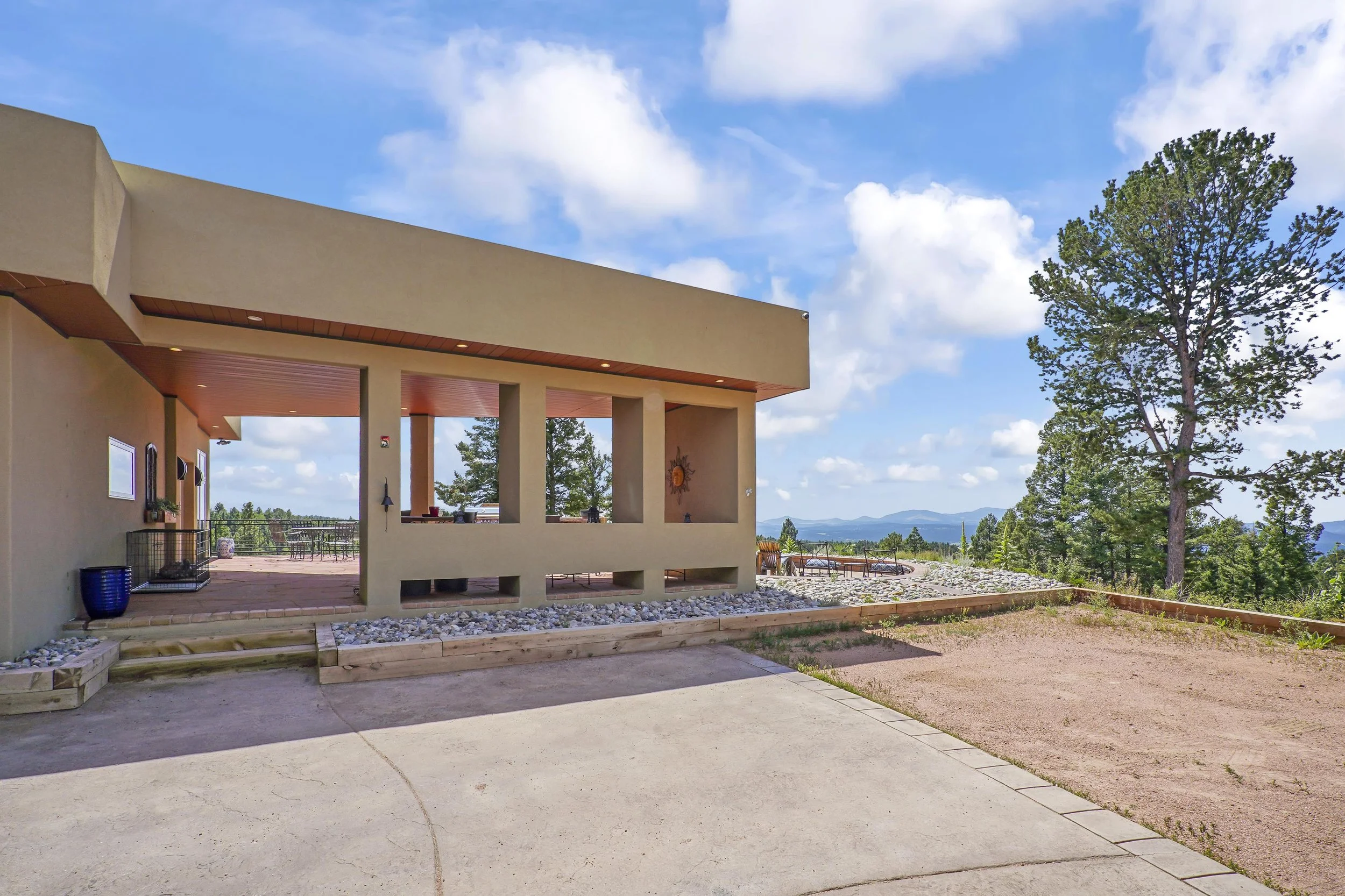 Modern house with a large outdoor patio, surrounded by trees and mountains in the distance under a partly cloudy sky.