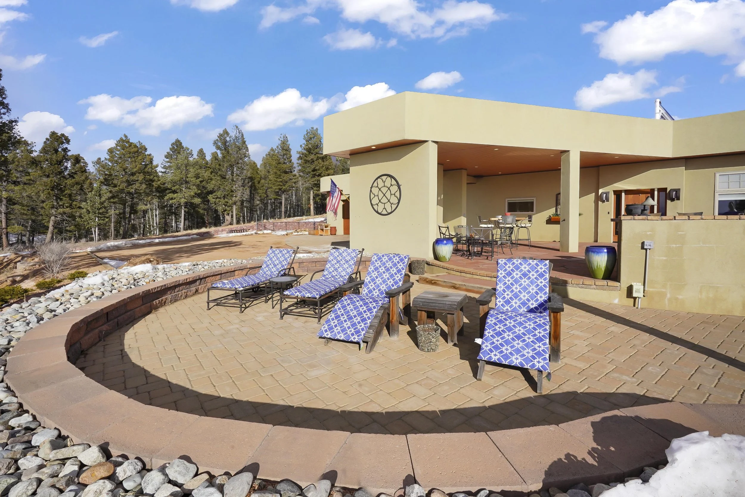 Outdoor patio with lounge chairs, tables, and potted plants in front of a modern house with a covered porch, surrounded by trees and a blue sky.