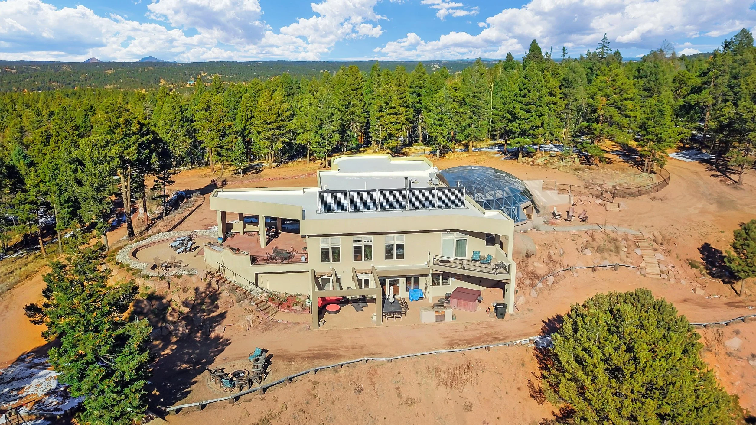 A multi-level house with a desert-like yard, surrounded by tall pine trees, under a partly cloudy sky, with mountains in the distance.
