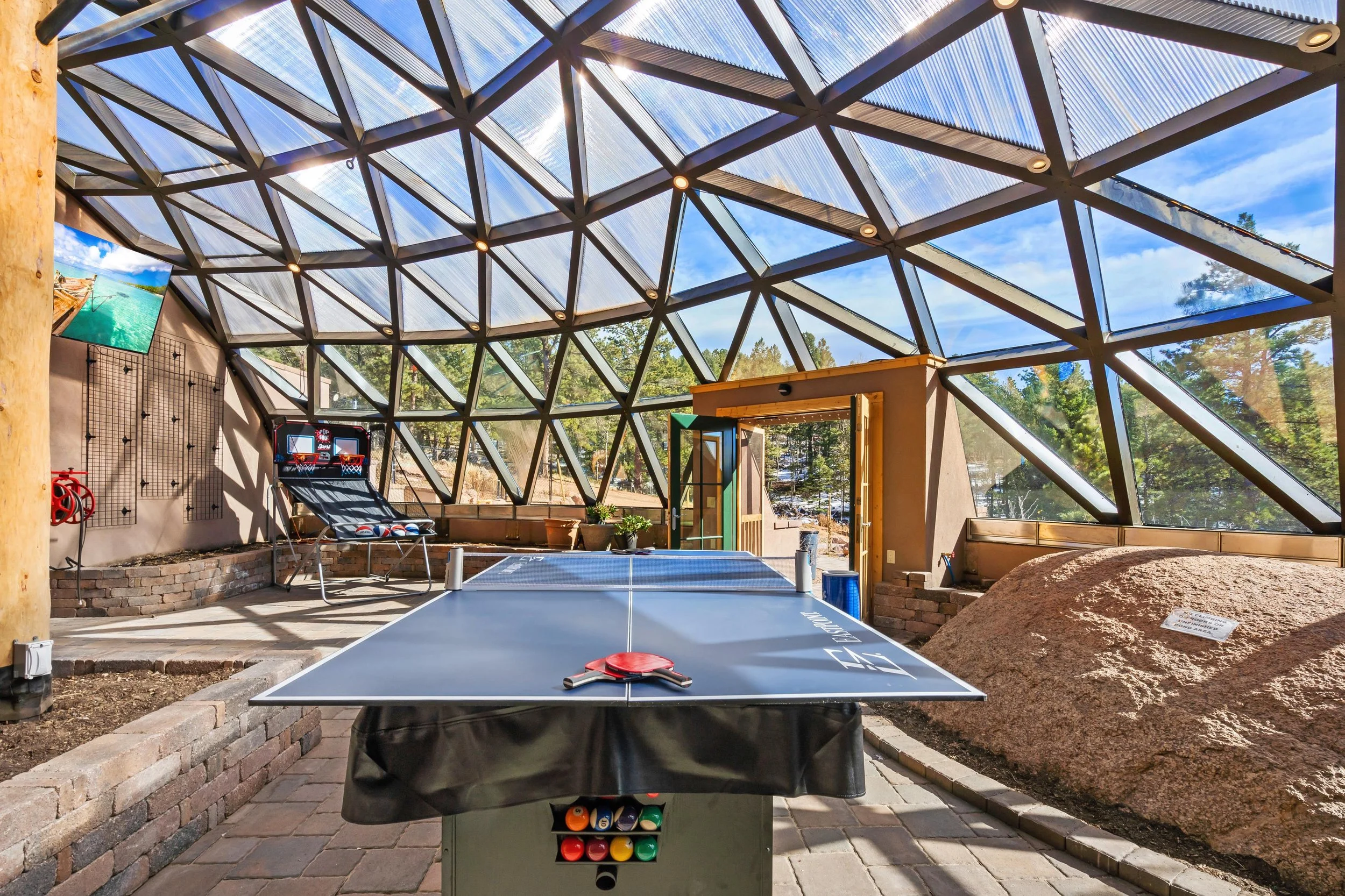 Indoor recreational space with a ping pong table in the foreground, a basketball arcade game in the background, a rock on the right, and large glass geometric dome ceiling allowing sunlight to stream in.
