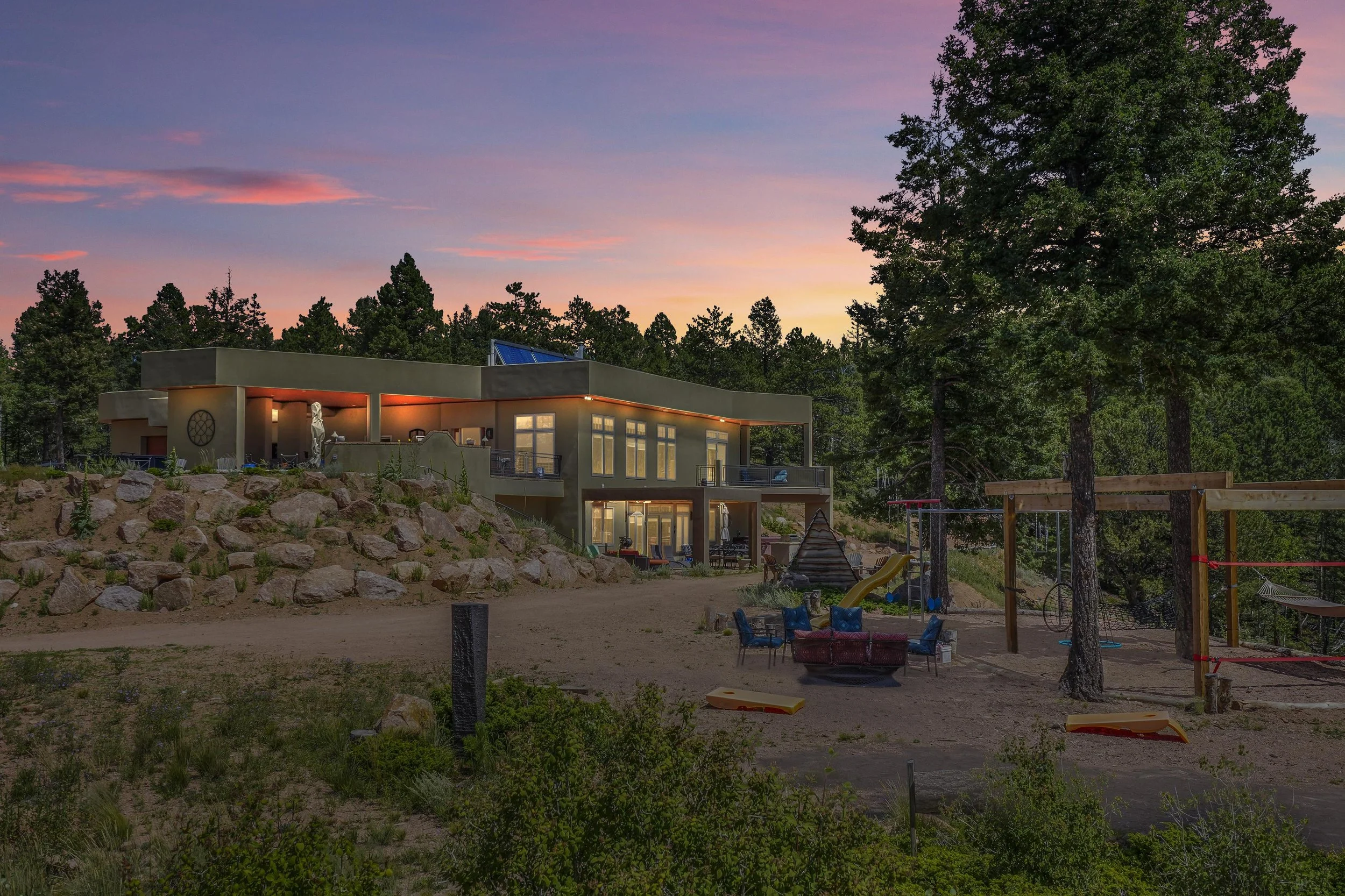 Modern house on a hillside surrounded by trees, with a backyard featuring a slide, swings, and outdoor furniture during sunset.