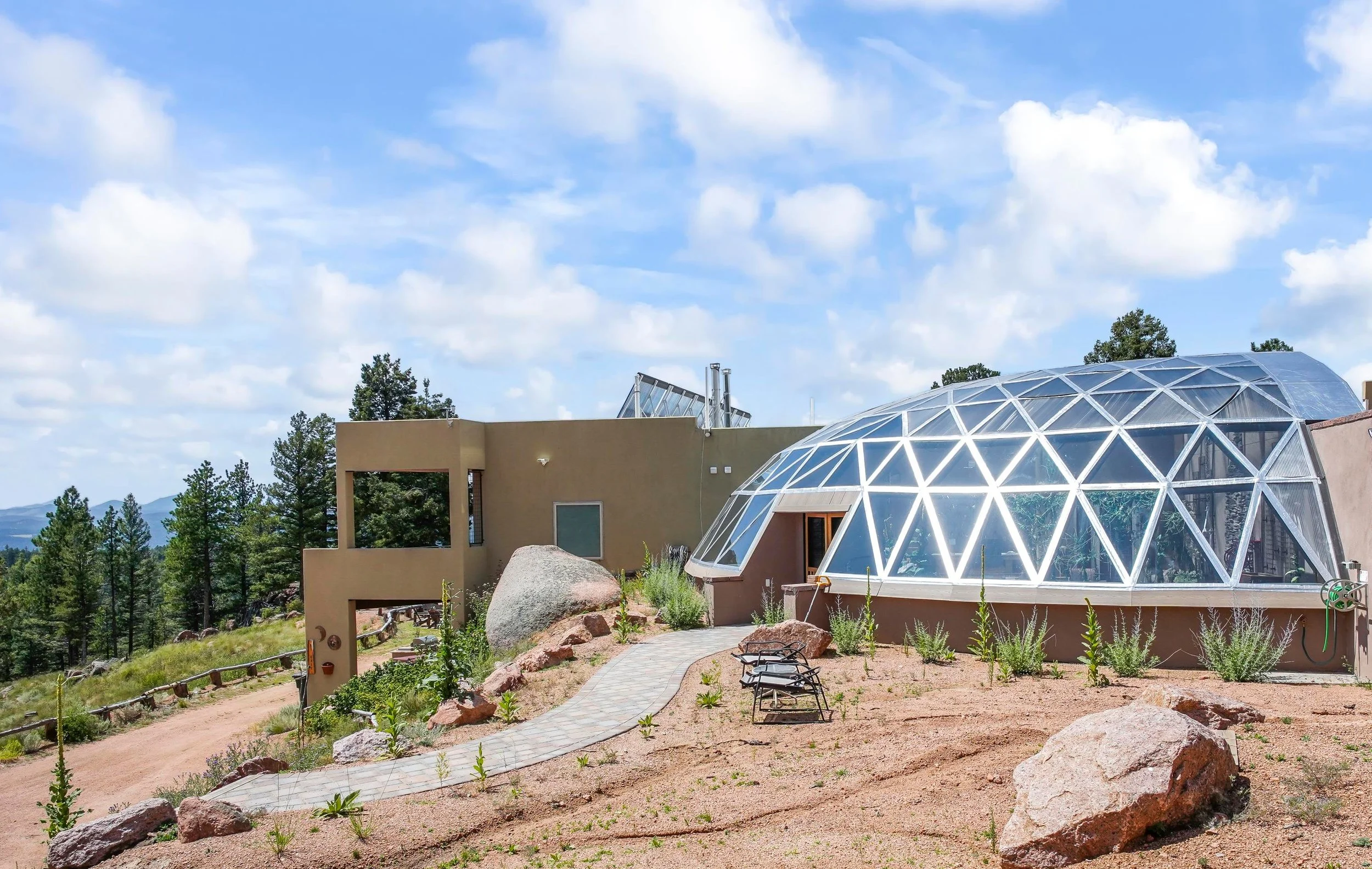Modern house with a large transparent geodesic dome structure on a hillside, surrounded by pine trees and a partly cloudy sky.