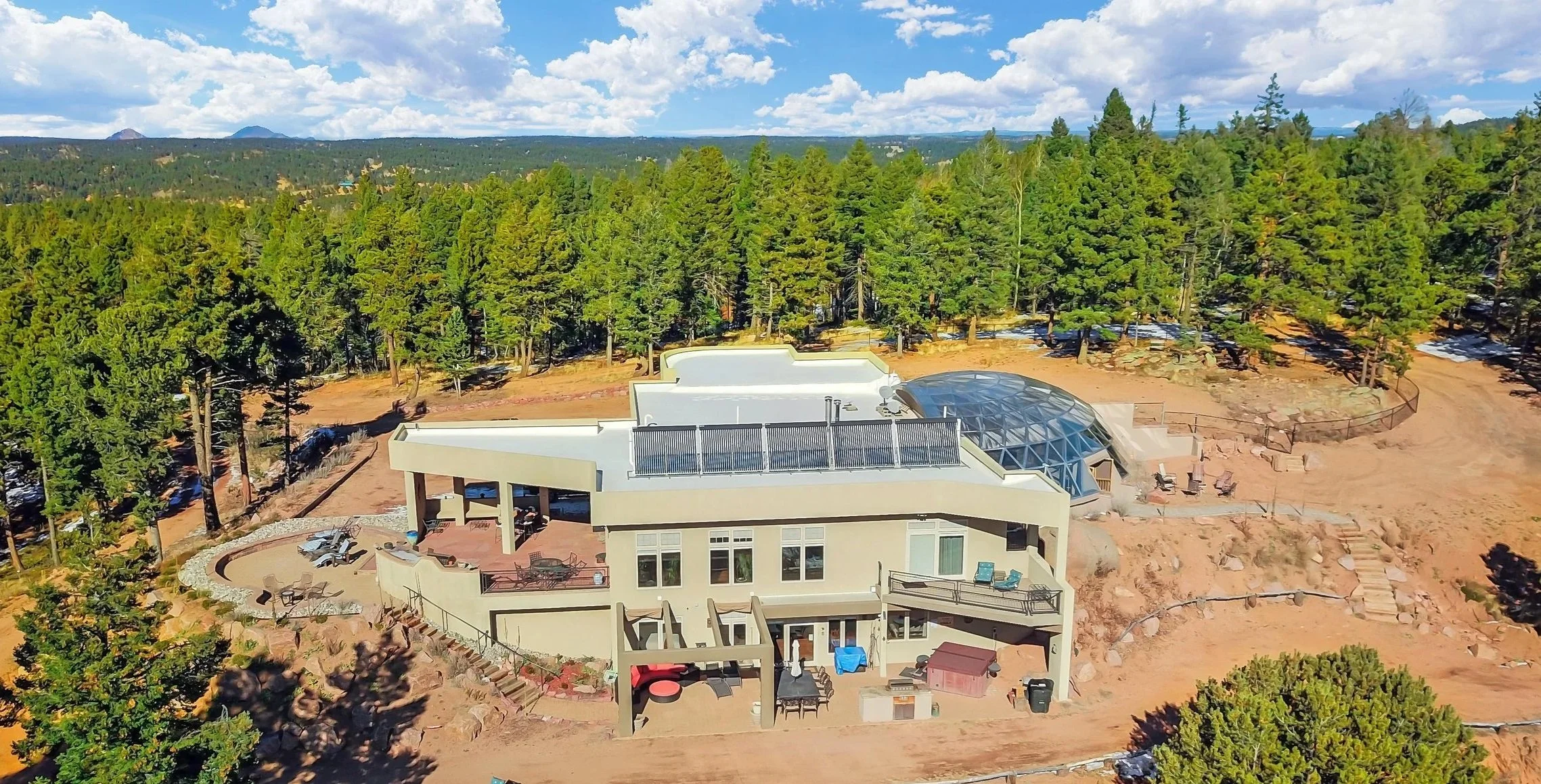 A modern multi-level house surrounded by trees on a hillside, with outdoor seating, a patio, and a glass-enclosed structure, under a blue sky with scattered clouds.
