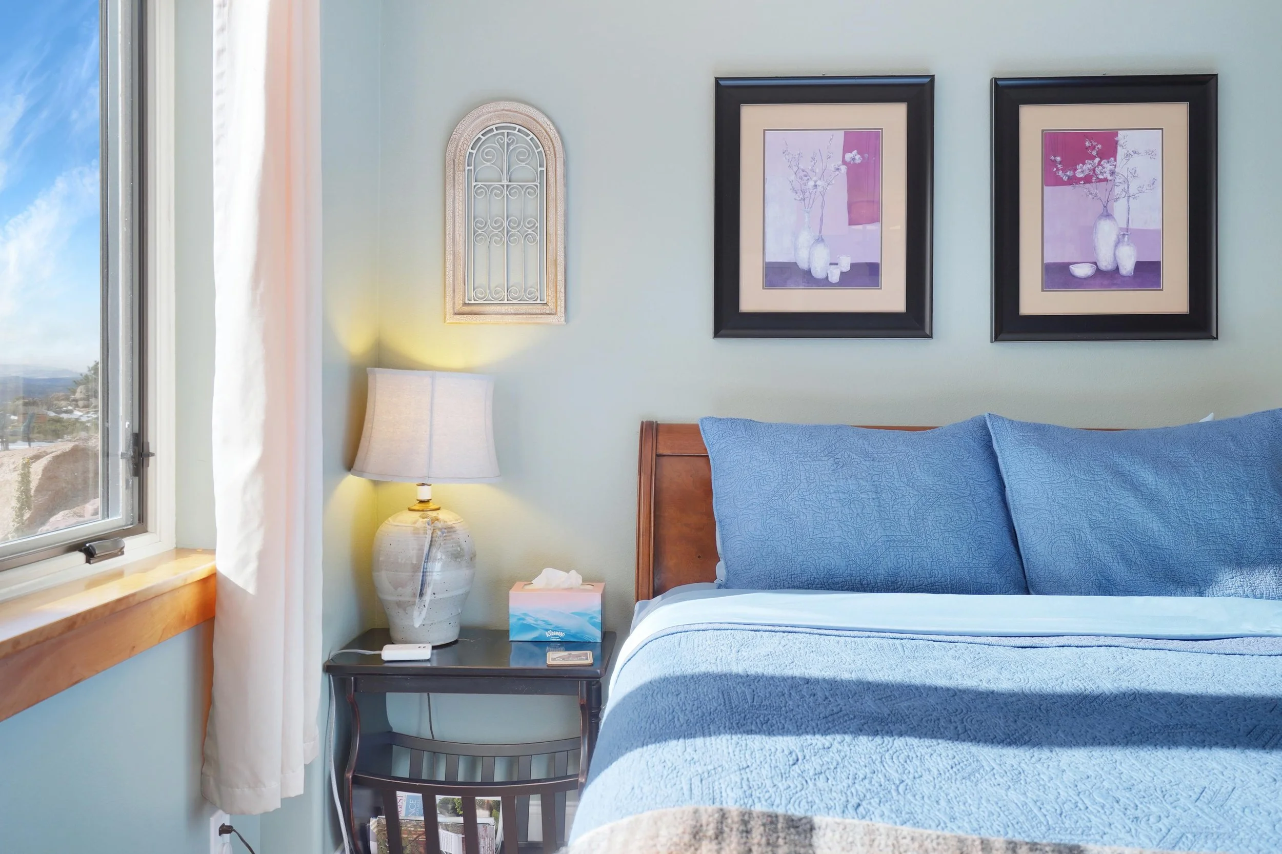 A bedroom with a window, white curtain, nightstand with a lamp, tissue box, and remote control, blue bed with pillows, framed artwork of vases with flowers on the wall.