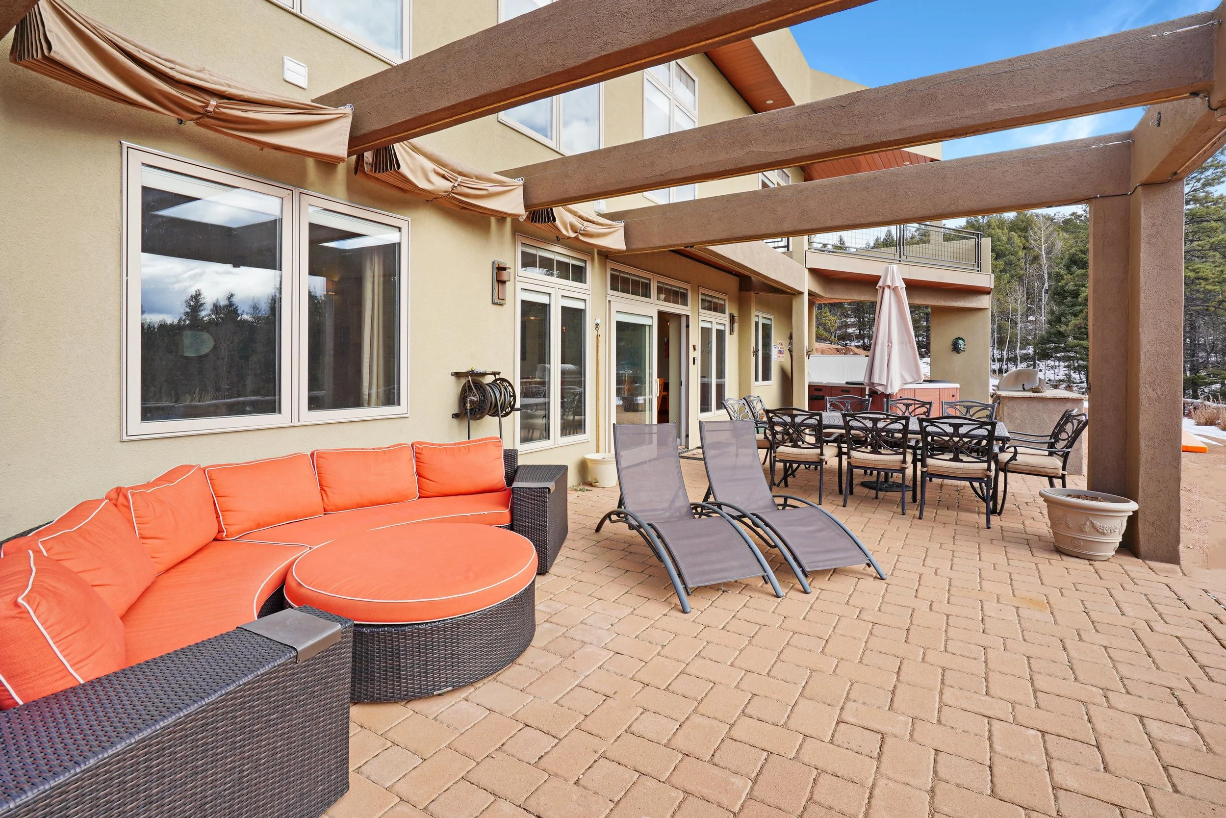 Outdoor patio with wicker sectional sofa with orange cushions, two gray lounge chairs, a dining table with eight chairs, and a hot tub in the background. The patio has a pergola with wooden beams and brick flooring, with snow visible in the backgroun