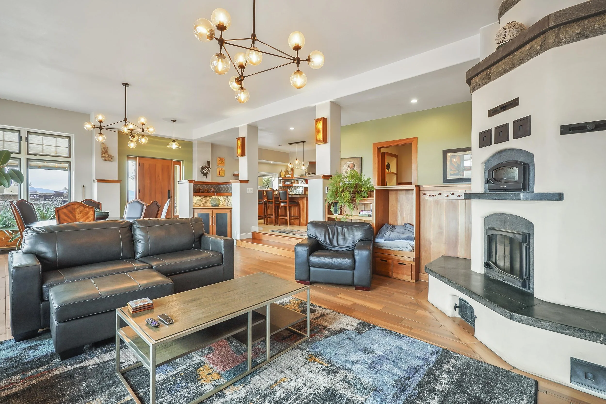 Open-concept living room with black leather couches, a wood coffee table, a fireplace, and a view into the dining and kitchen areas with pendant lighting and wooden accents.