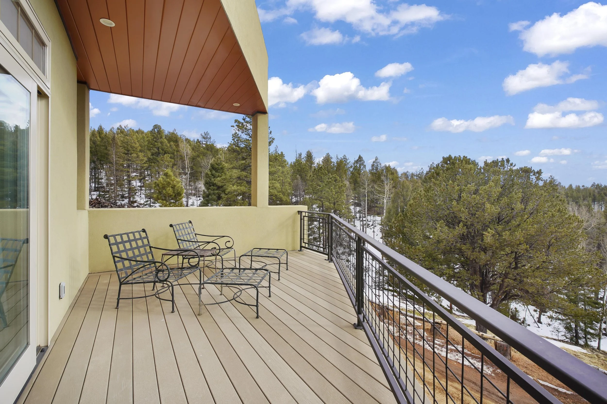 Balcony with four black metal chairs and a small table, overlooking a snowy forested landscape with pine trees under a partly cloudy blue sky.