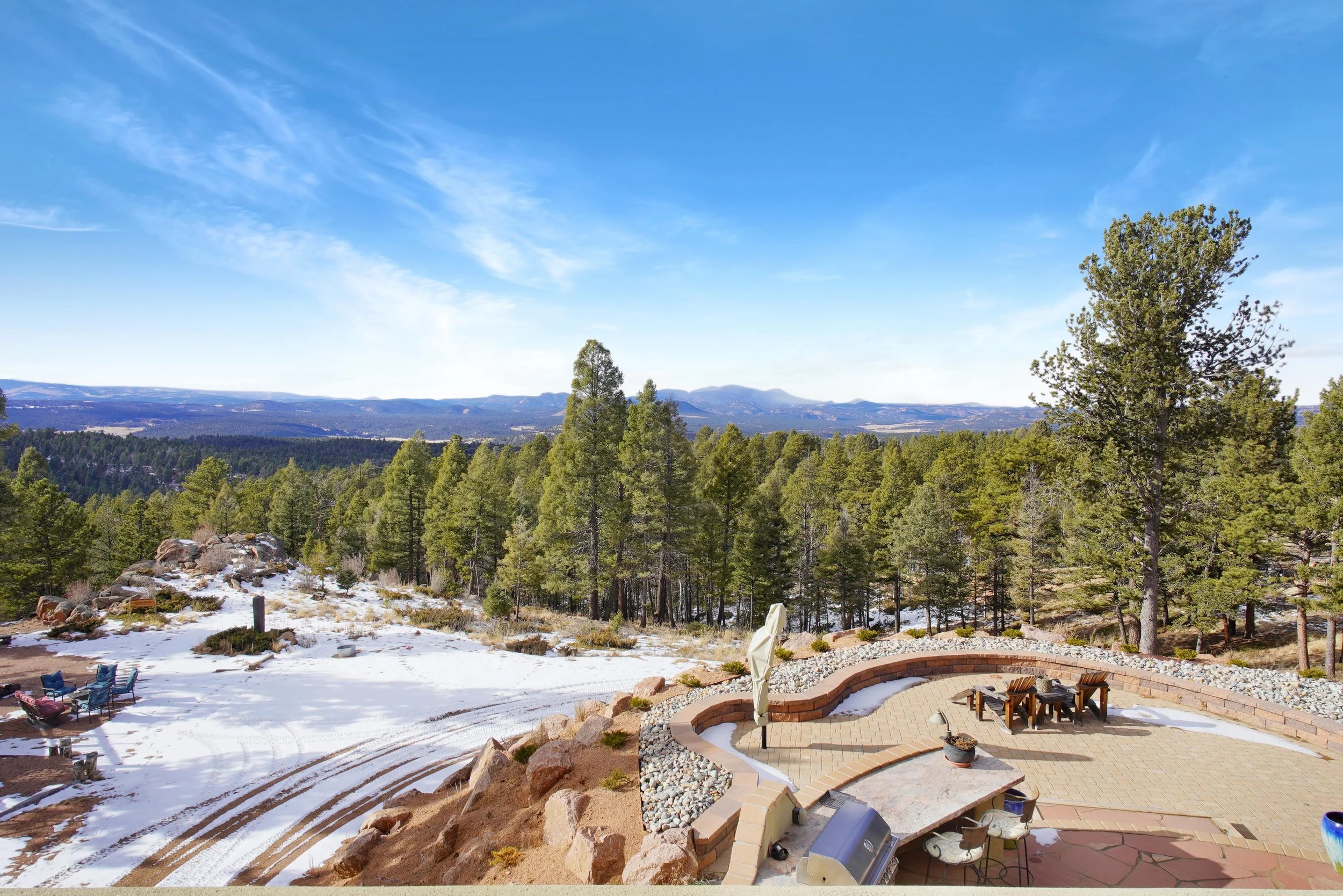 A scenic outdoor patio with a brick and stone design, surrounded by a natural landscape of pine trees and distant mountains. The patio includes a table, chairs, and an umbrella, with some snow on the ground and tire tracks in a nearby dirt area.