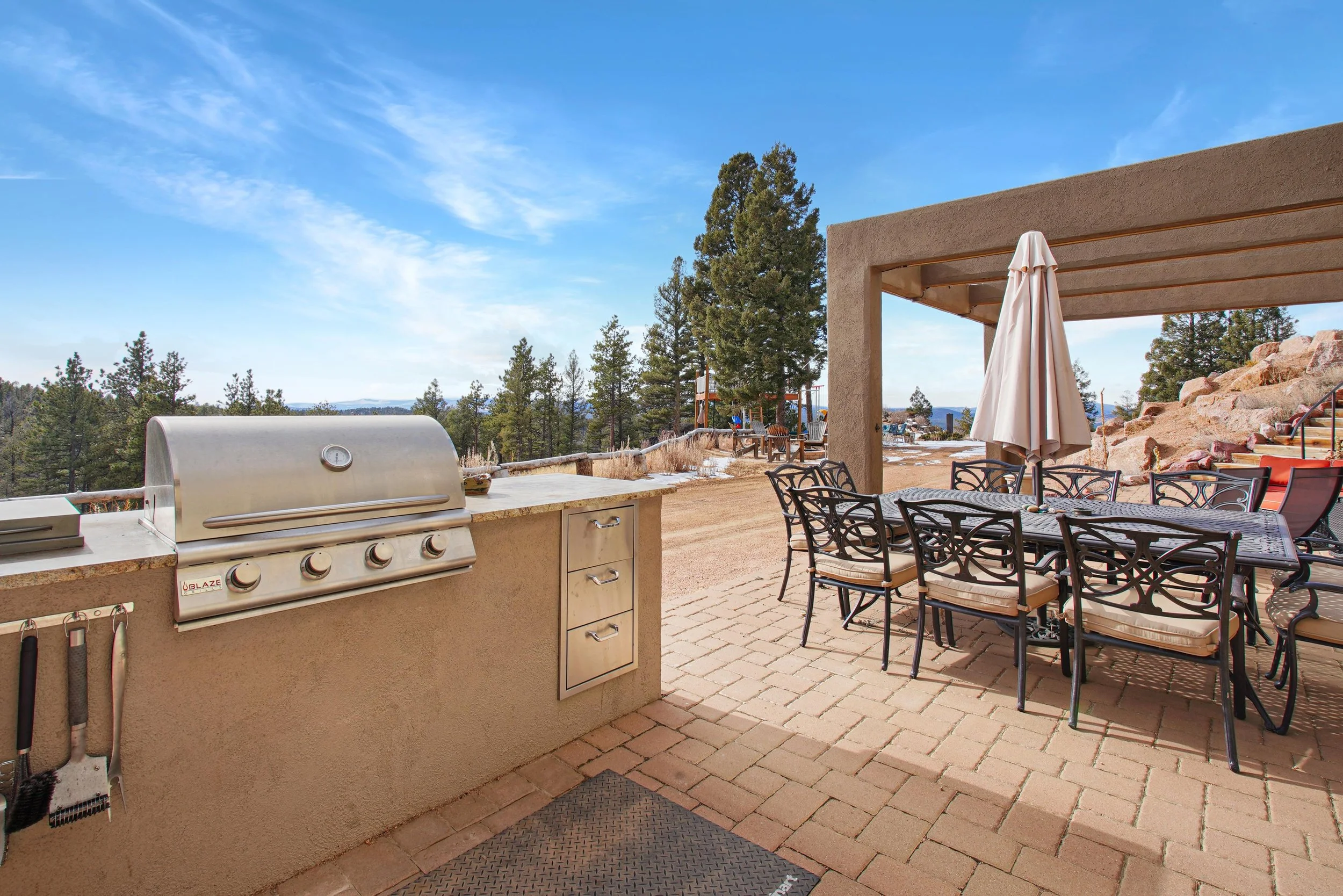 Outdoor patio with a built-in grill, a dining table with chairs and an umbrella, against a backdrop of trees and clear blue sky.
