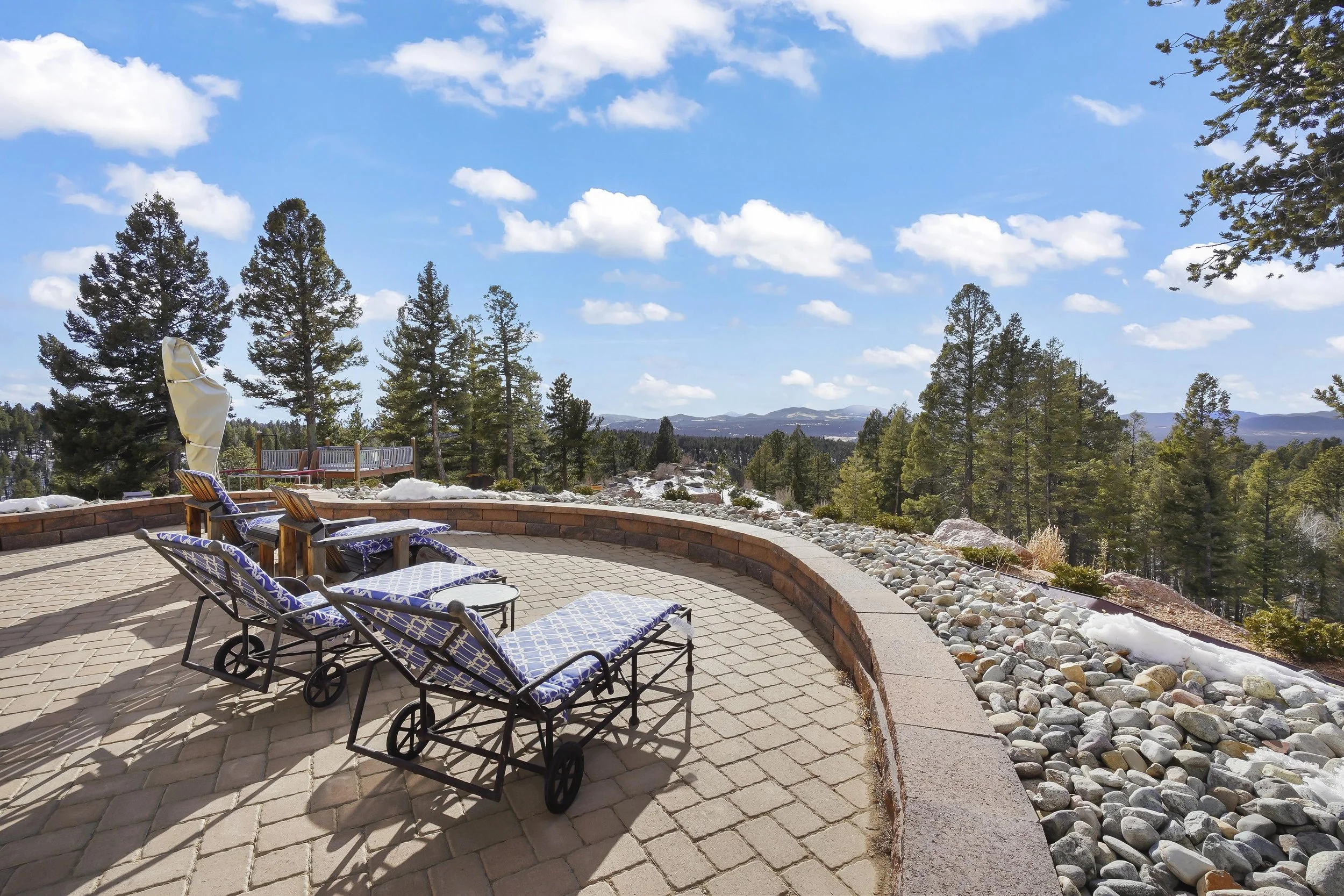 A spacious outdoor patio with lounge chairs and a small table, surrounded by a low brick wall and a rocky landscape, overlooking pine trees and distant mountains under a blue sky with white clouds.