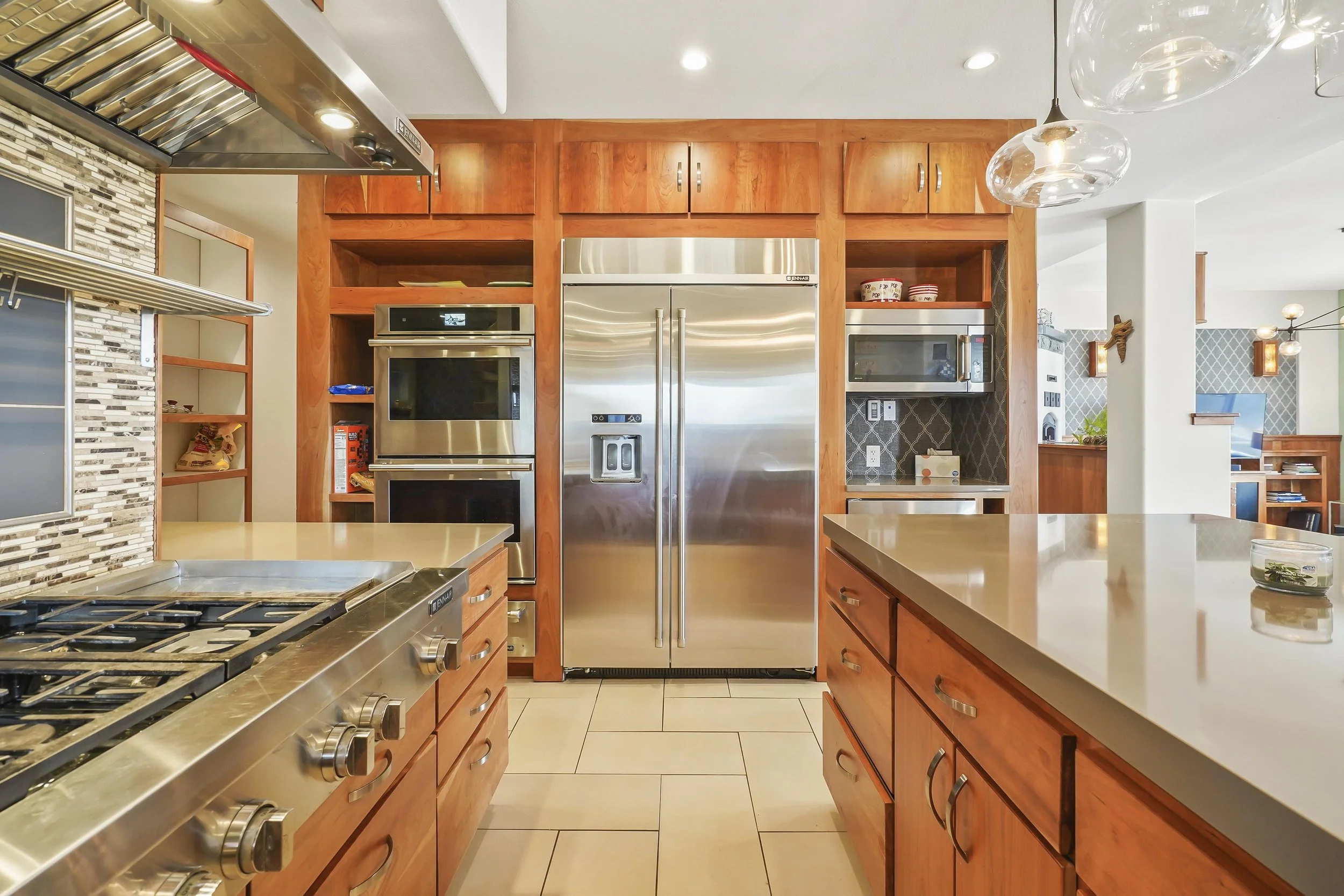 Modern kitchen with stainless steel refrigerator, double ovens, microwave, gas stovetop, wooden cabinets, glass pendant lights, and tiled backsplash.