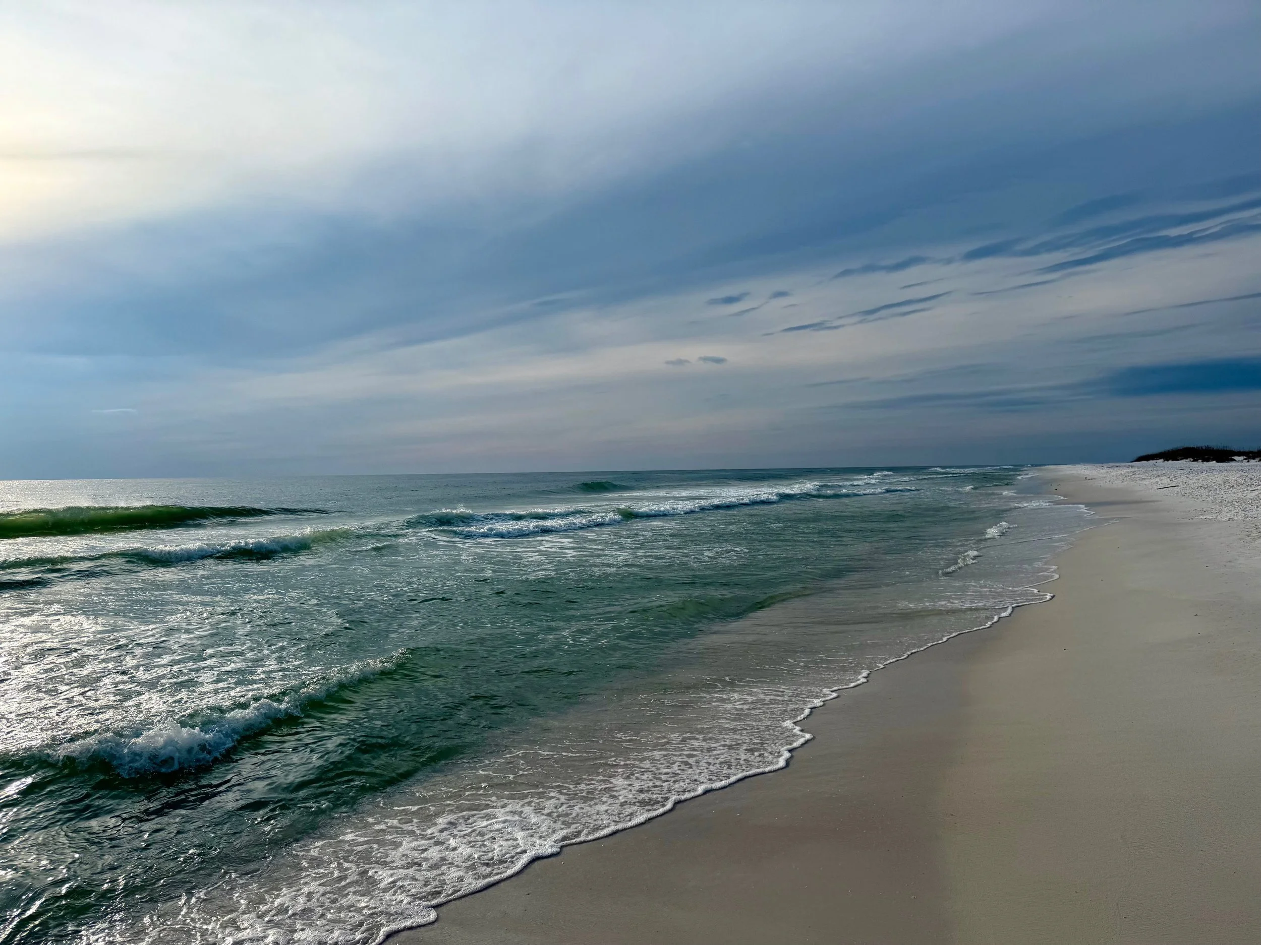 Empty beach with white sand, gentle waves, and a cloudy sky over the ocean.