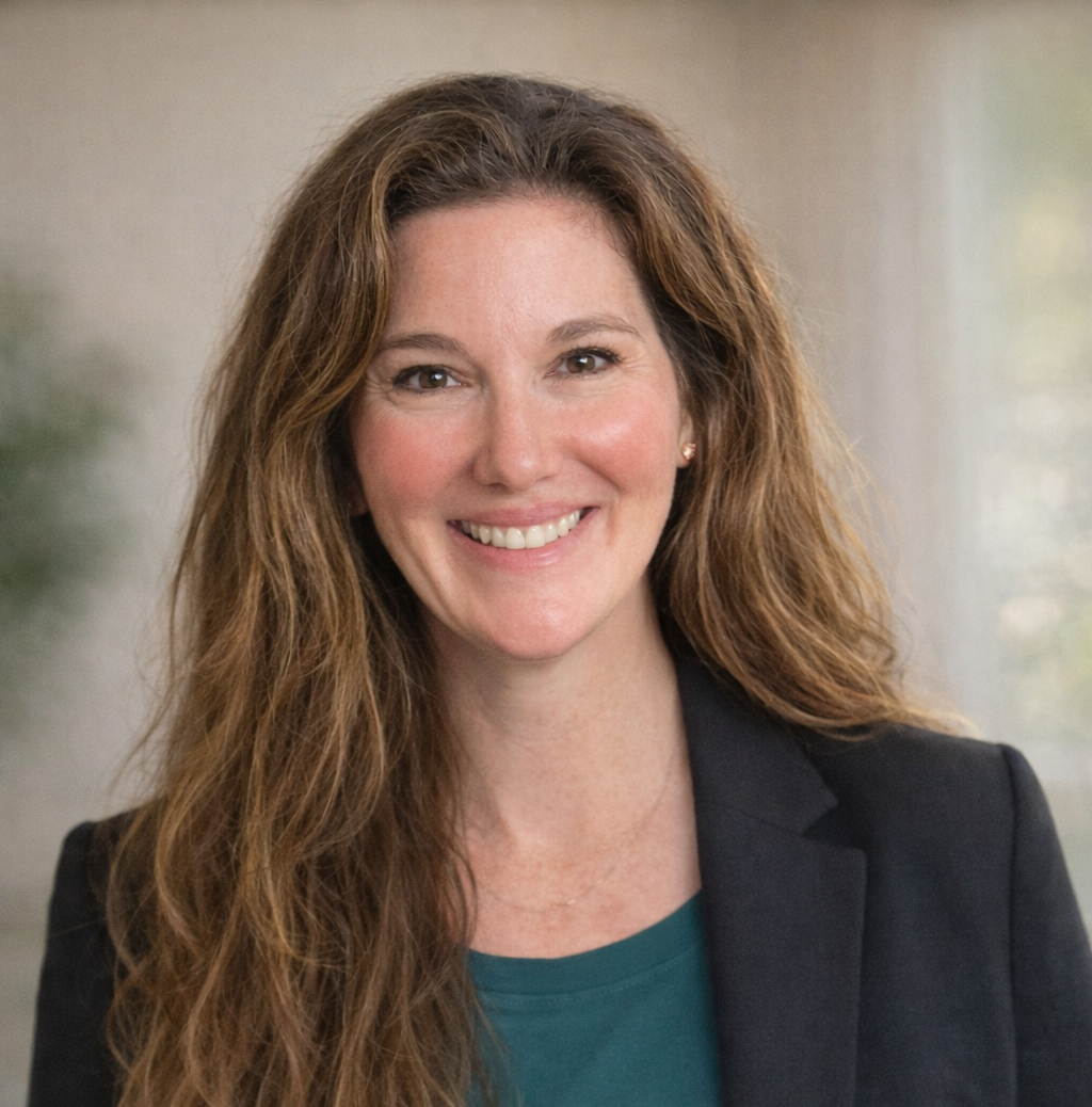 A woman with long, curly brown hair, wearing a dark blazer and a teal shirt, smiling in an indoor setting with a blurred background.