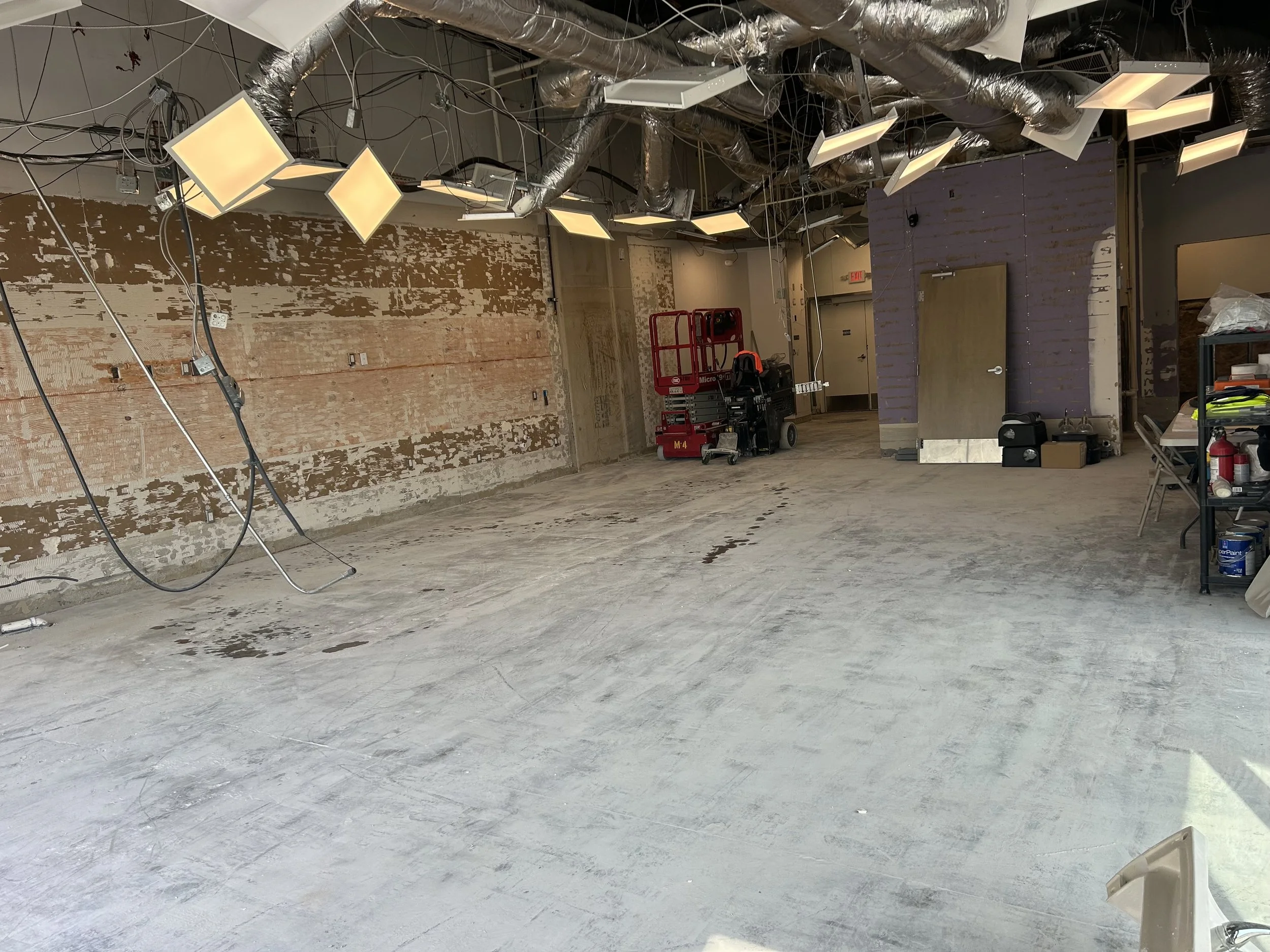 Empty commercial space under renovation with unfinished walls, exposed ductwork, lighting fixtures hanging from the ceiling, and construction equipment and supplies scattered around.