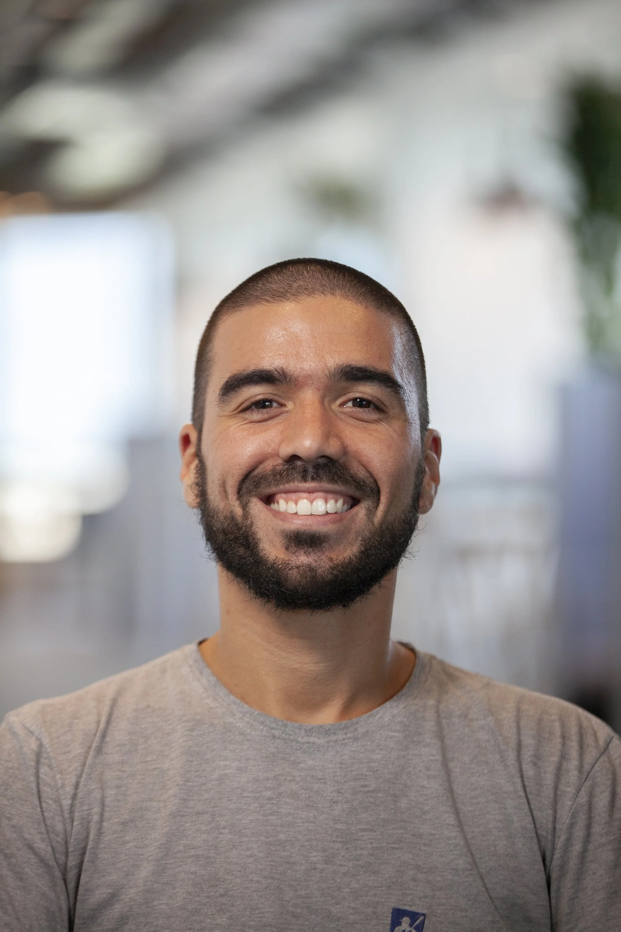 Homem de pele clara, cabelo curto e barba sorrindo, usando camiseta cinza, em ambiente bem iluminado com fundo desfocado de cores neutras e verde.