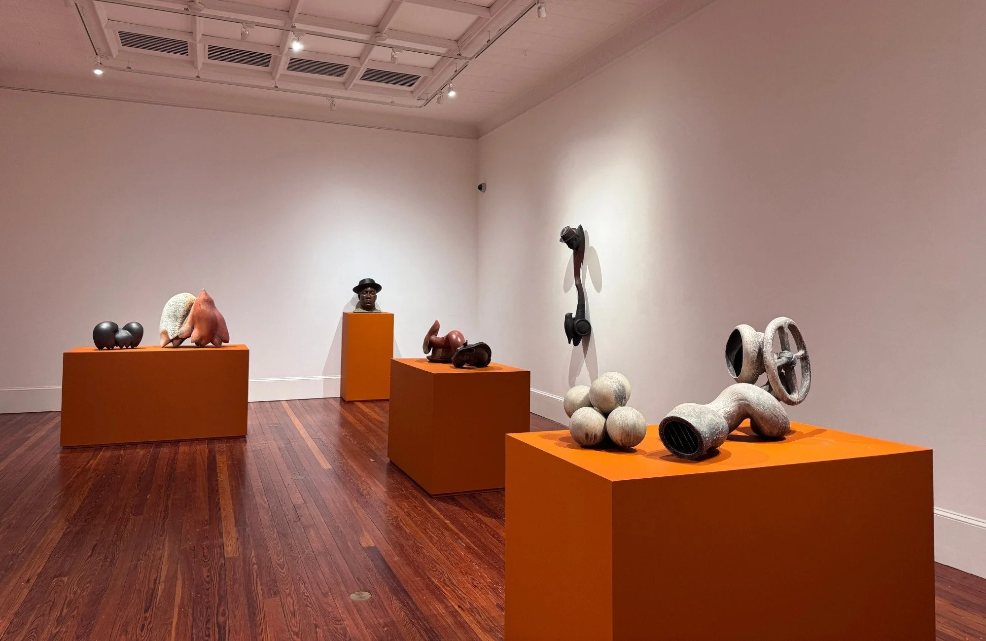 Art gallery with sculptures on orange pedestals, including abstract figures, a bust of a person with a hat, and industrial-style objects, set against white walls and a wooden floor.