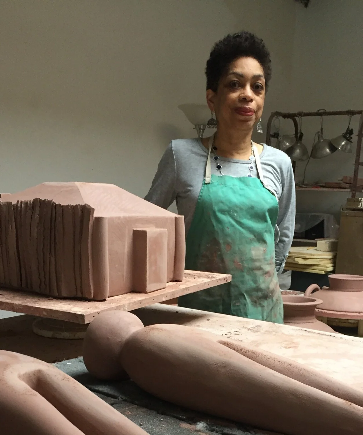 Artist, Syd Carpenter, wearing a green apron and gray shirt standing behind pottery sculptures in a studio.