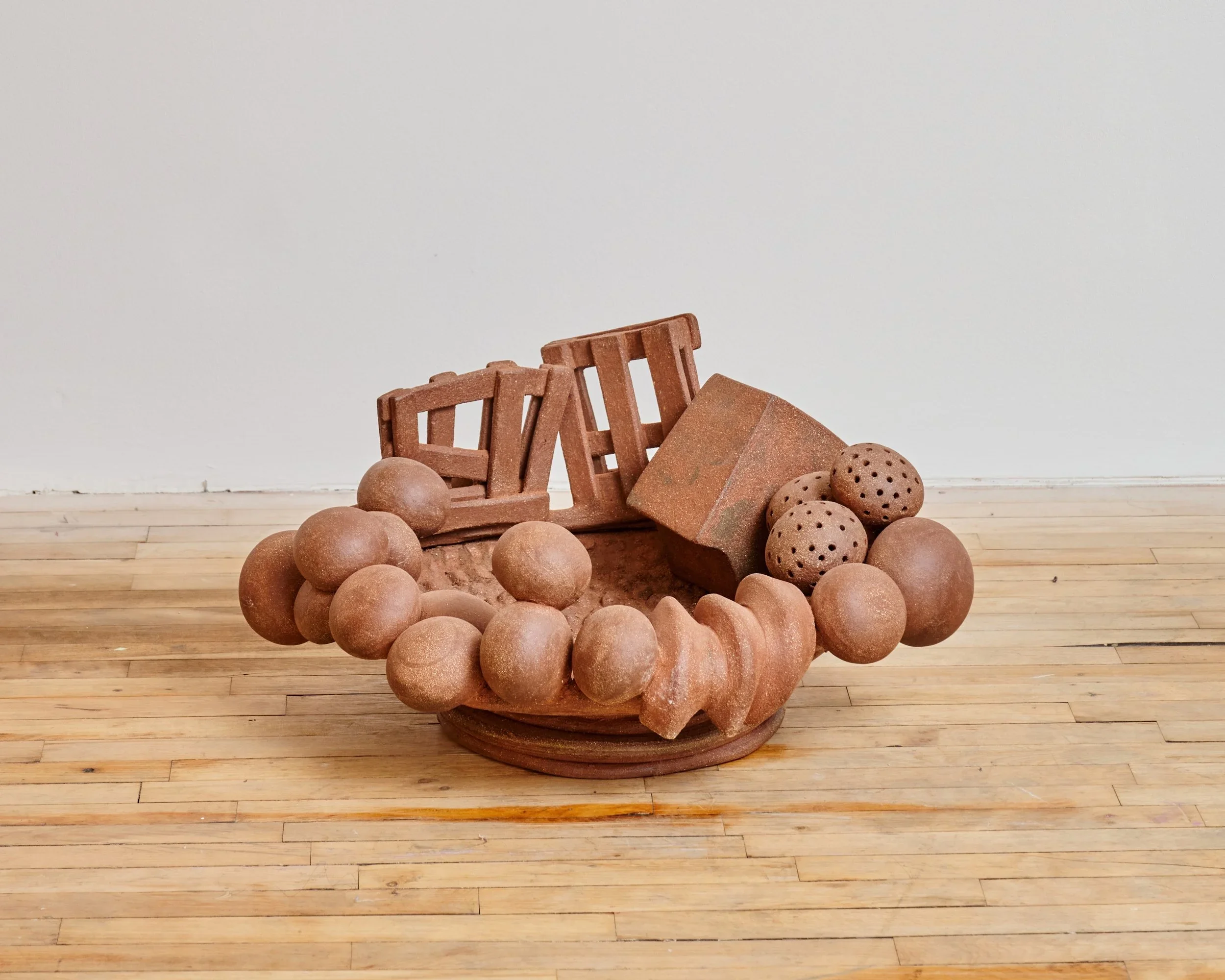 Clay sculpture of a sofa with round armrests, surrounded by clay balls and geometric shapes on a wooden floor against a plain wall.