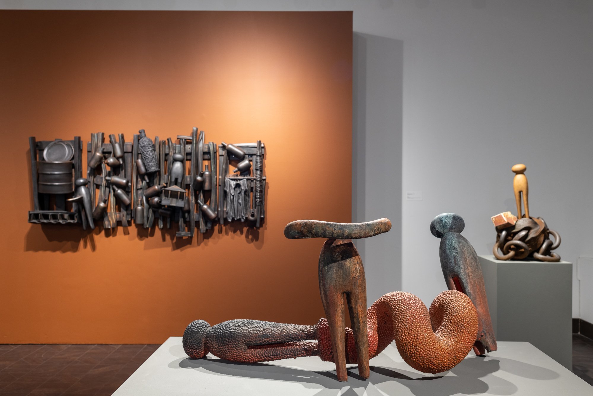 Museum exhibit featuring wooden sculptures, a textured chair, and stone objects, with a wall-mounted metal art piece and orange and gray walls in the background.