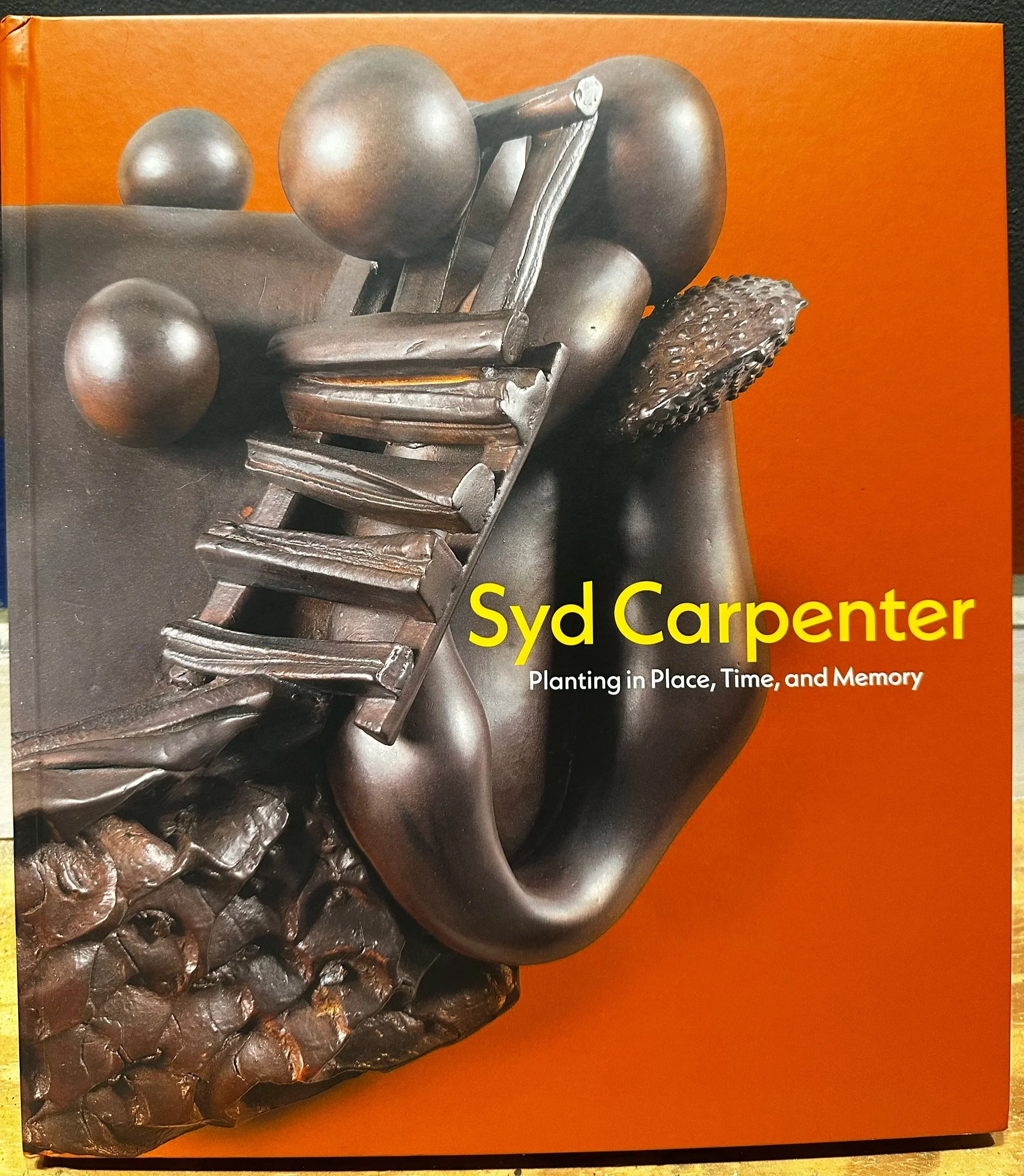 Book cover featuring a metallic sculpture of a person with balls and a textured object, orange background, yellow title text.