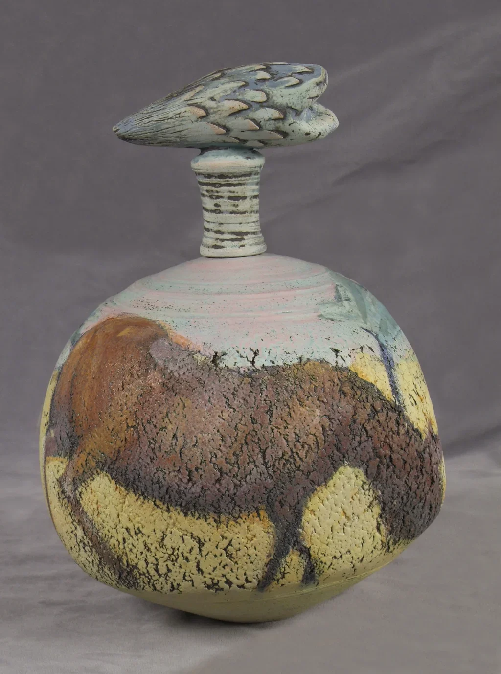 A ceramic sculpture of a bison painted with earthy tones, featuring a rounded body and a stylized head on a cylindrical neck, set against a plain grey background.