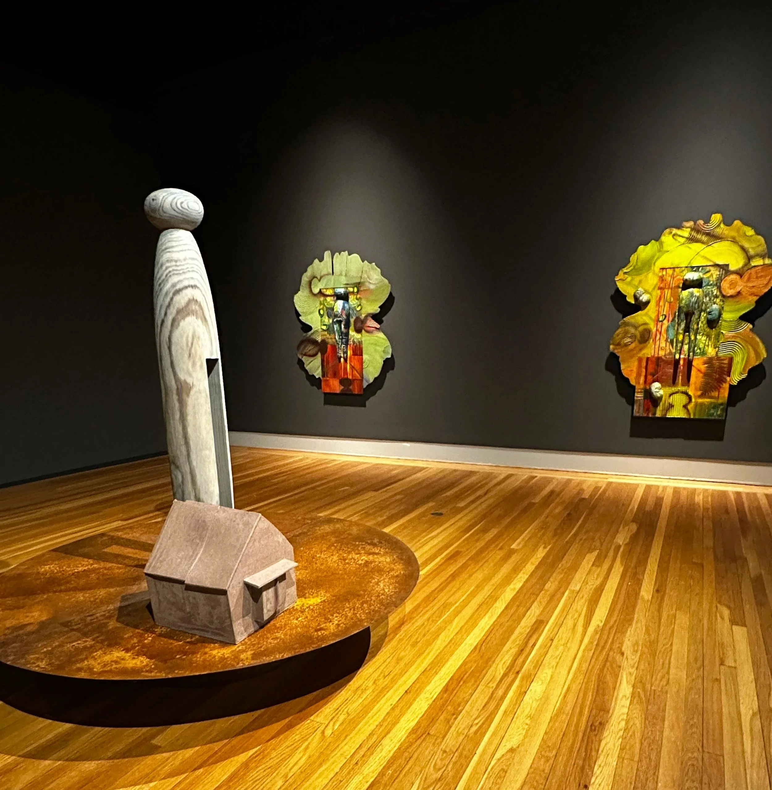 Wooden exhibit table with a grey stone sculpture, 4 colorful abstract sculptures with intricate designs on a dark gallery wall with warm wood flooring.