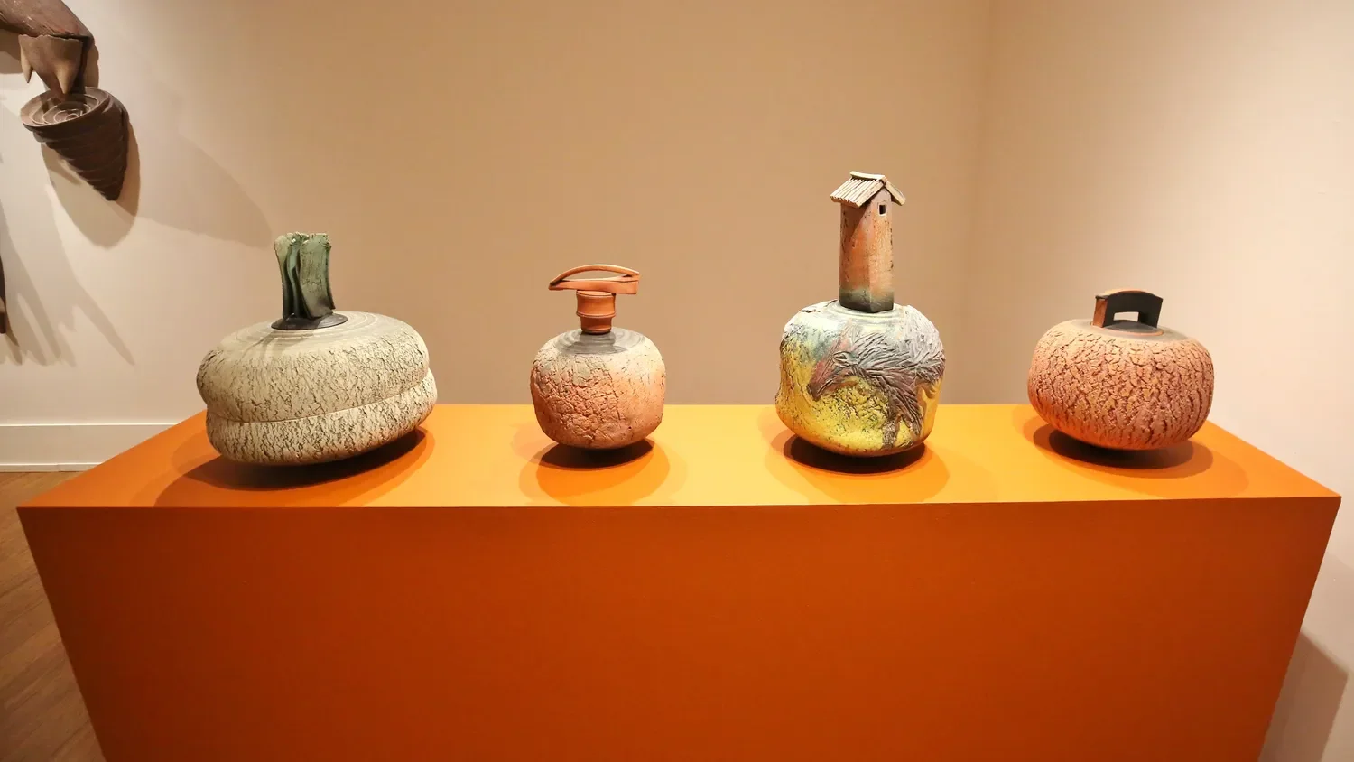 Four ceramic art sculptures on an orange display table in a gallery. Each piece is unique with textured surfaces and different shapes, including pots with handles and a tall, narrow sculpture with a small roof.