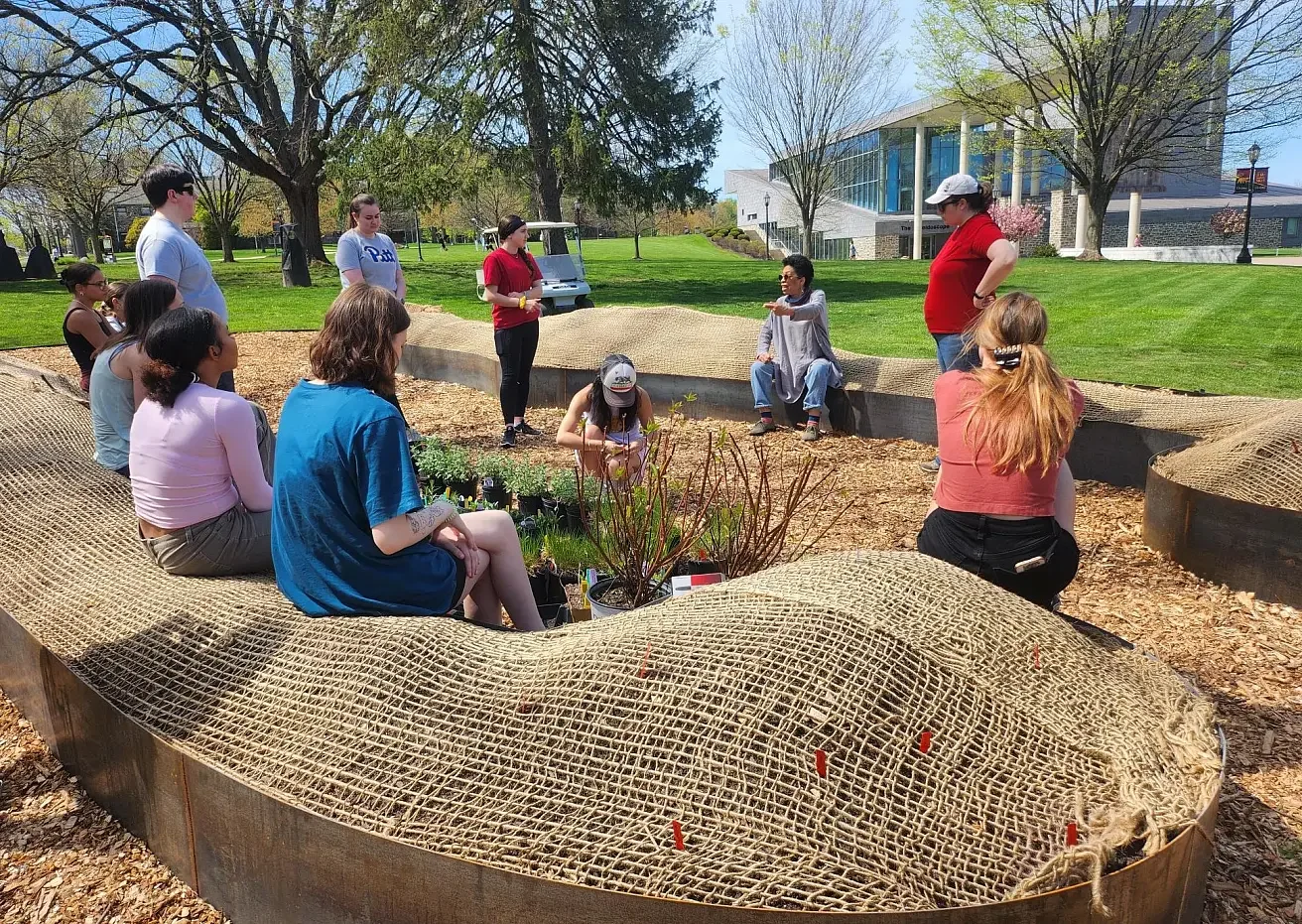 A group of people gathered outdoors around a garden bed, with some sitting and others standing, during a sunny day with trees and a modern building in the background.