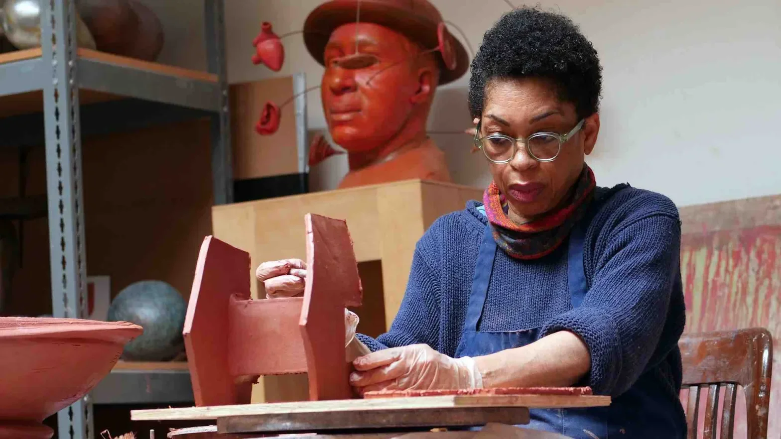 Syd Carpenter working with clay in a pottery studio, with sculptures and ceramic objects in the background.