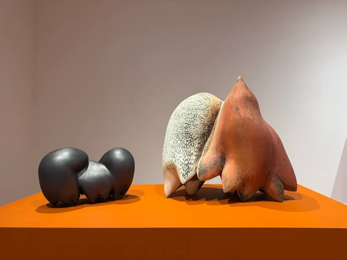 Two abstract sculptures on an orange surface. The sculpture on the left is small, black, rounded, and smooth. The sculpture on the right is larger, irregularly shaped, with a textured surface and a reddish-brown color.