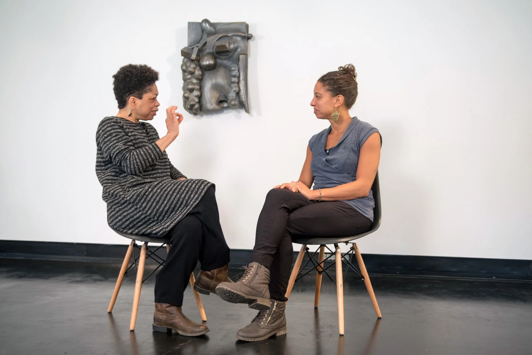 Two women sitting on chairs facing each other, engaged in conversation, with a large abstract face sculpture on the white wall behind them, in a minimalist room with black floor and baseboard.