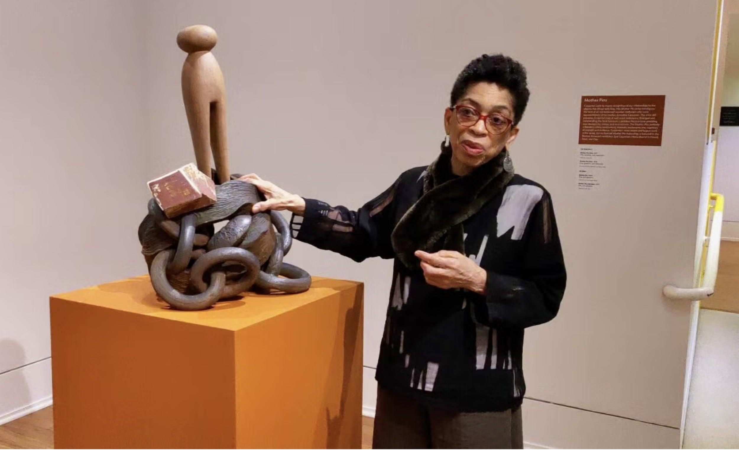 Artist Syd Carpenter wearing red glasses and a patterned blazer touching an abstract sculpture made of intertwined metallic loops on an orange pedestal at an art gallery.
