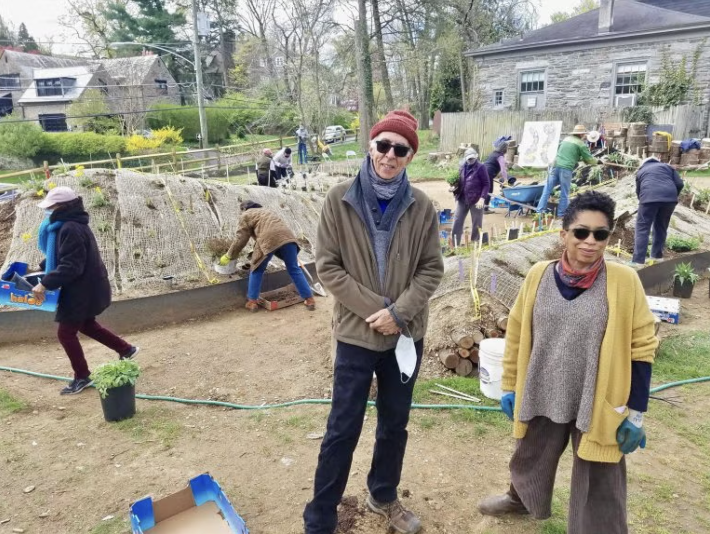 People working together on a community garden, planting and tending to the soil, with two individuals in the foreground wearing glasses and jackets, while others in the background are engaged in gardening activities.